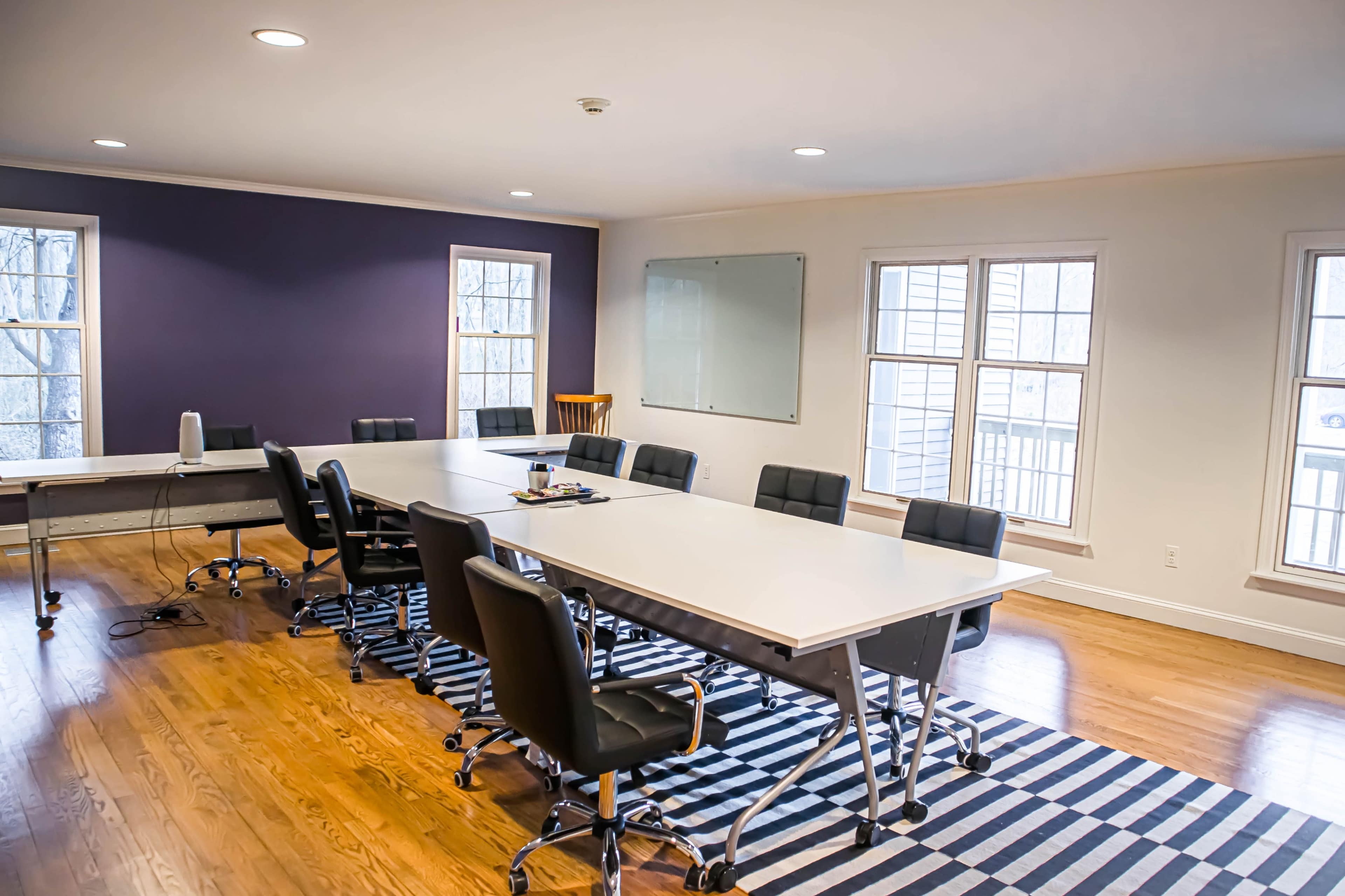 A spacious conference room features a long table surrounded by black rolling chairs, with large windows allowing natural light to illuminate the space.