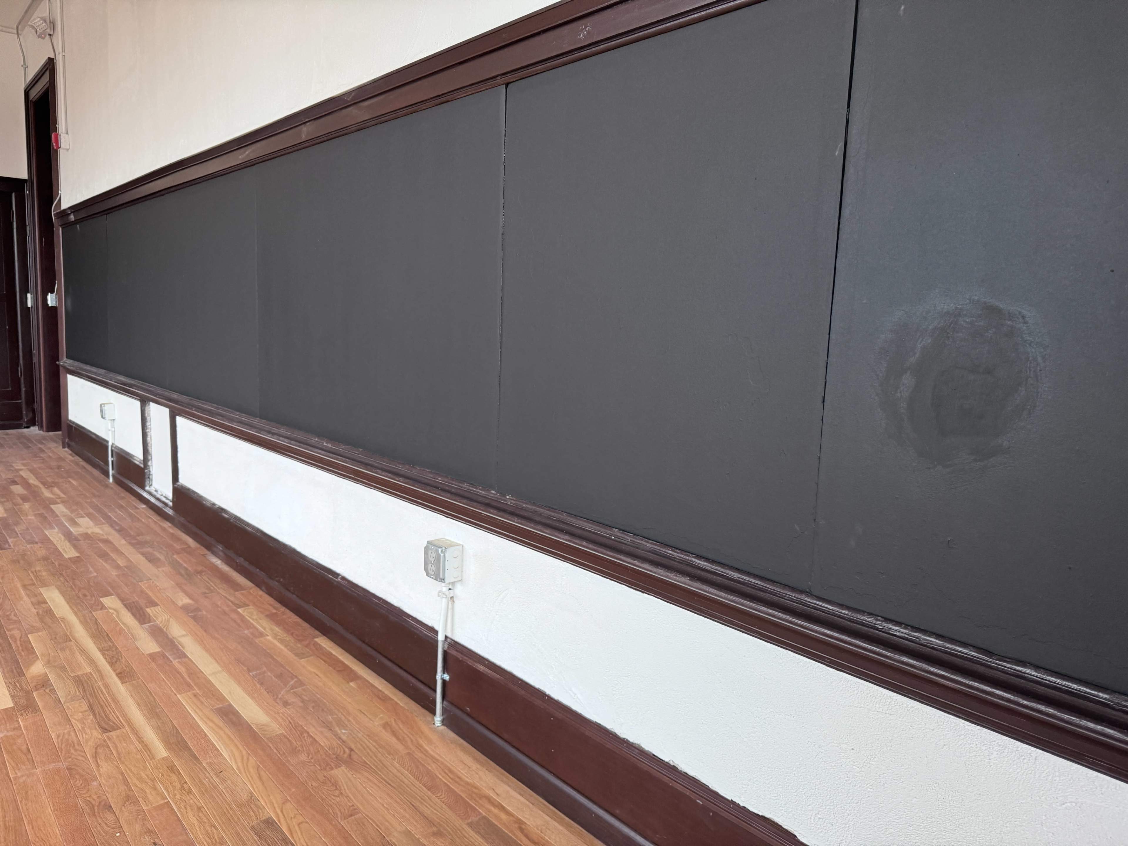 The image shows a wall with a blackboard surface, framed by dark wood trim, and a section of hardwood flooring beneath.