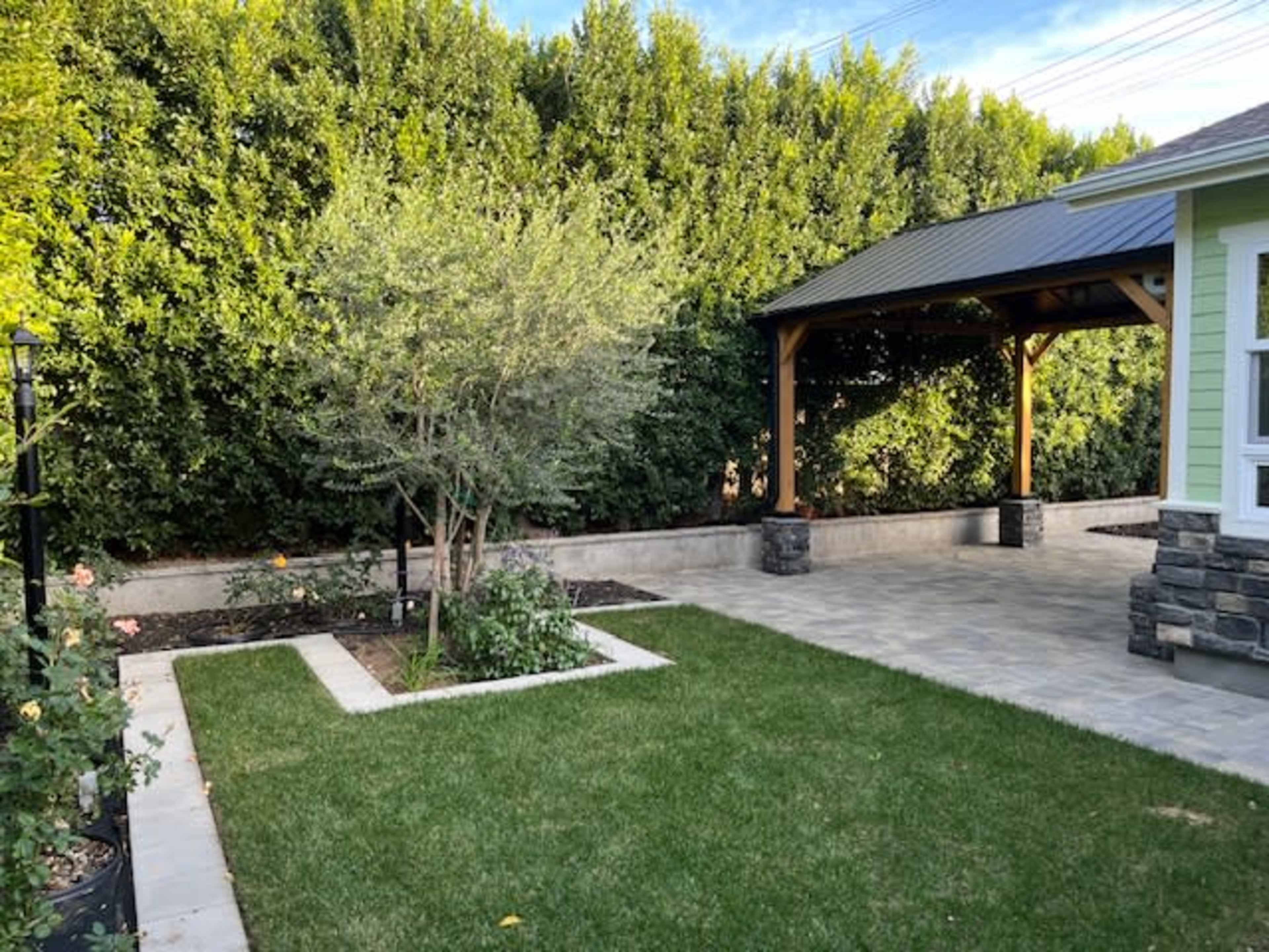 The image shows a landscaped backyard with a stone walkway, raised garden beds, and a covered patio surrounded by tall green shrubs.