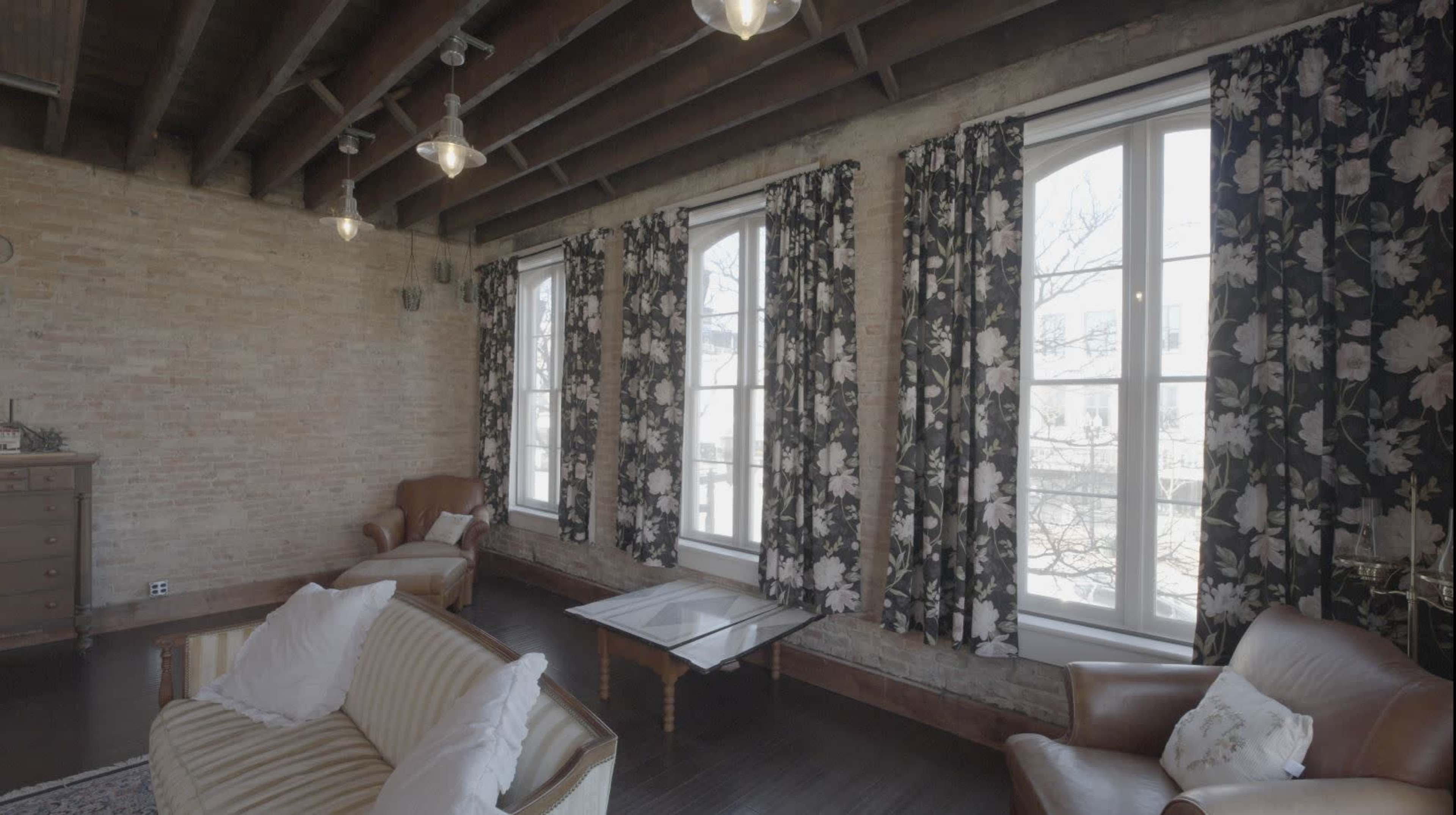 The image depicts a spacious room with large windows dressed in floral curtains, featuring two seating areas that include a patterned sofa and a leather armchair.