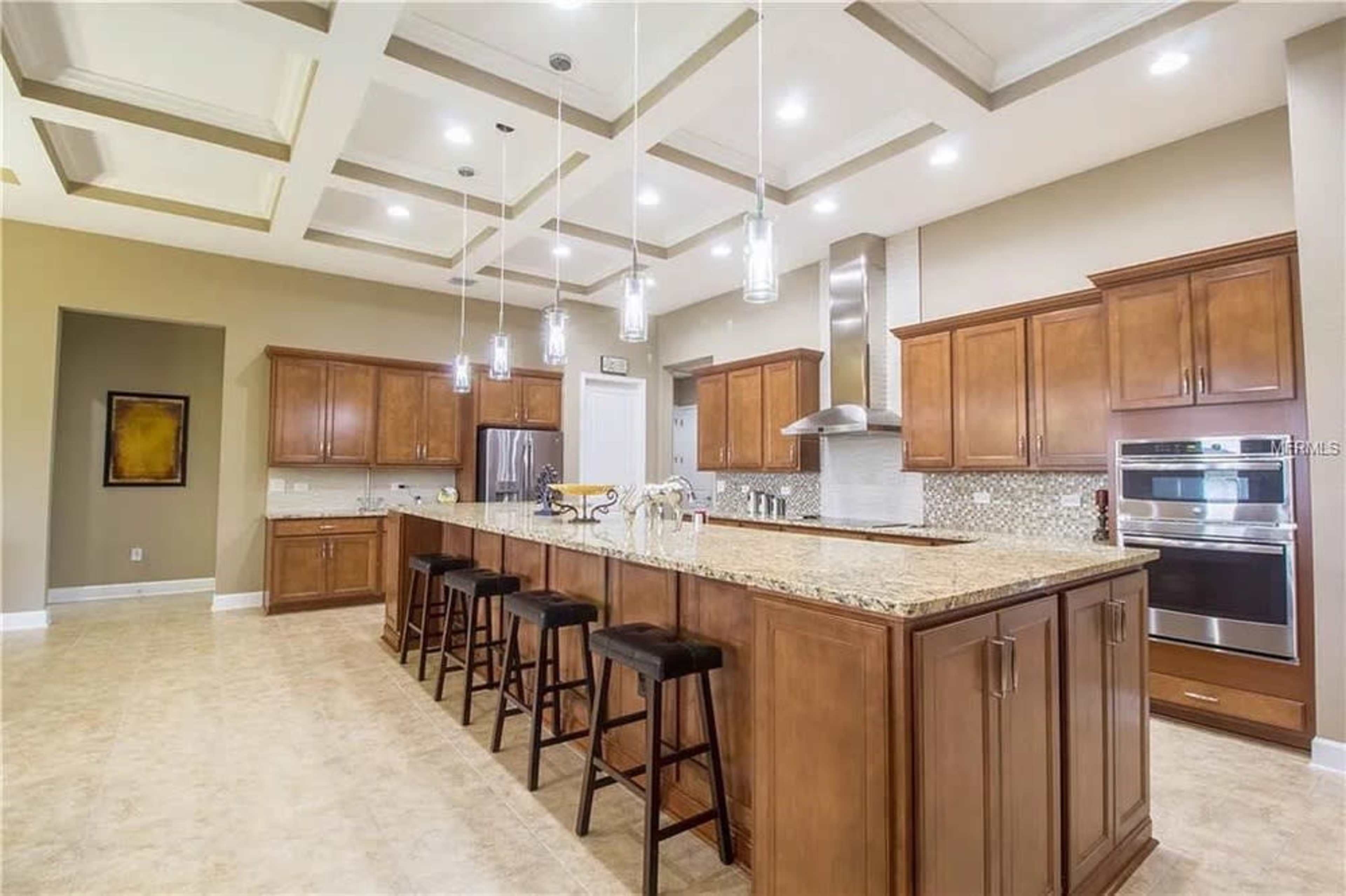 The image shows a spacious modern kitchen with wooden cabinetry, a large island with bar stools, and pendant lighting.
