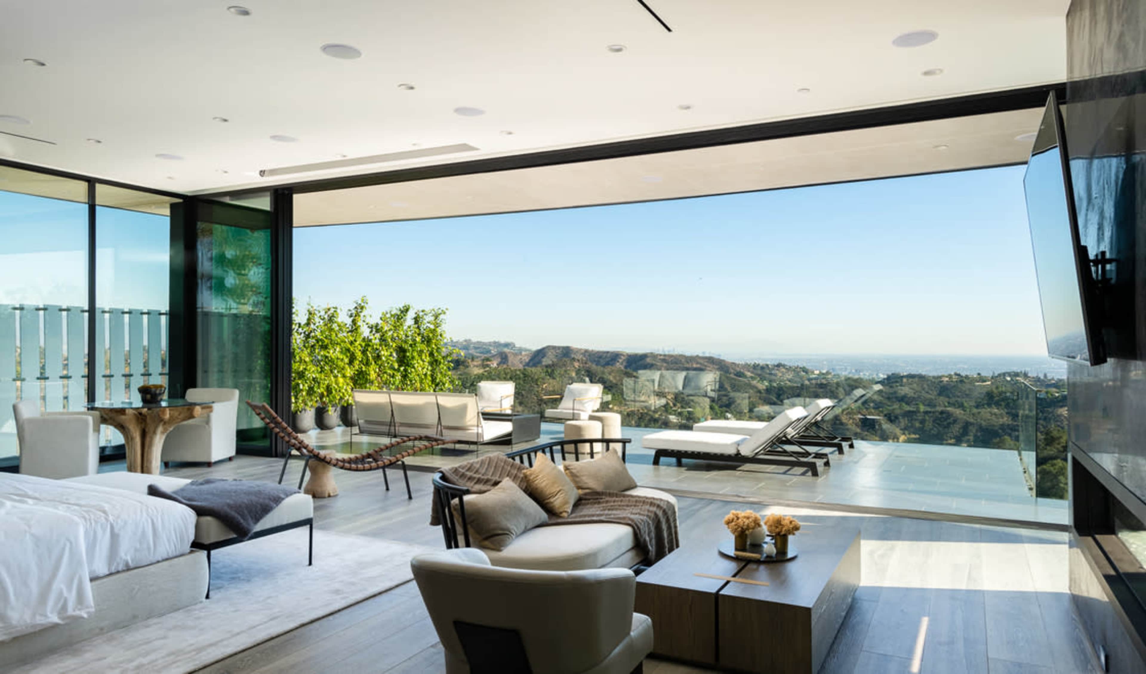 A spacious modern living room features floor-to-ceiling windows with a view of the hills, minimalist furniture, and a seamless transition to an outdoor terrace.