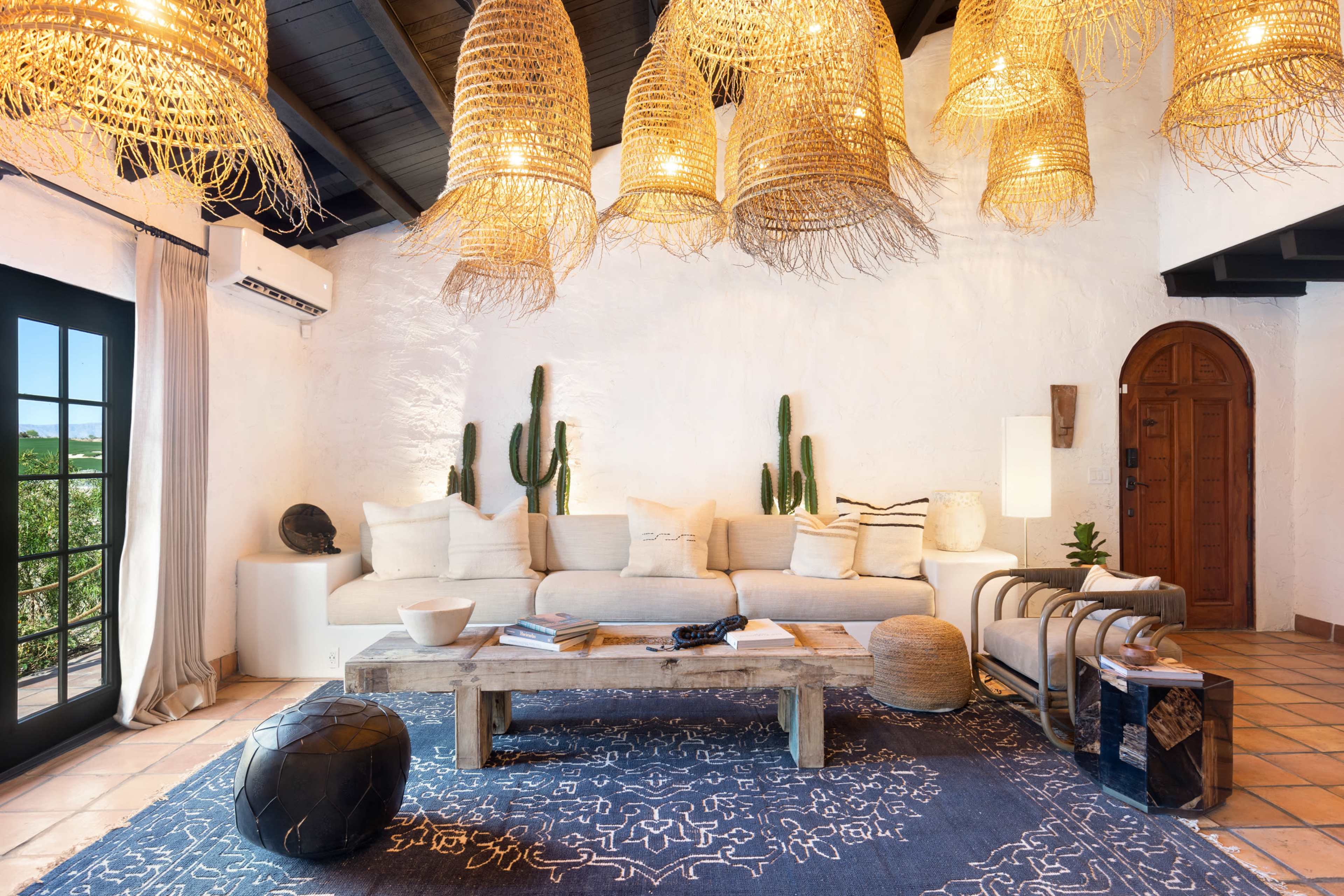The image shows a living room with a white-walled interior, featuring a large sofa with neutral cushions, hanging woven light fixtures, and decorative cacti.