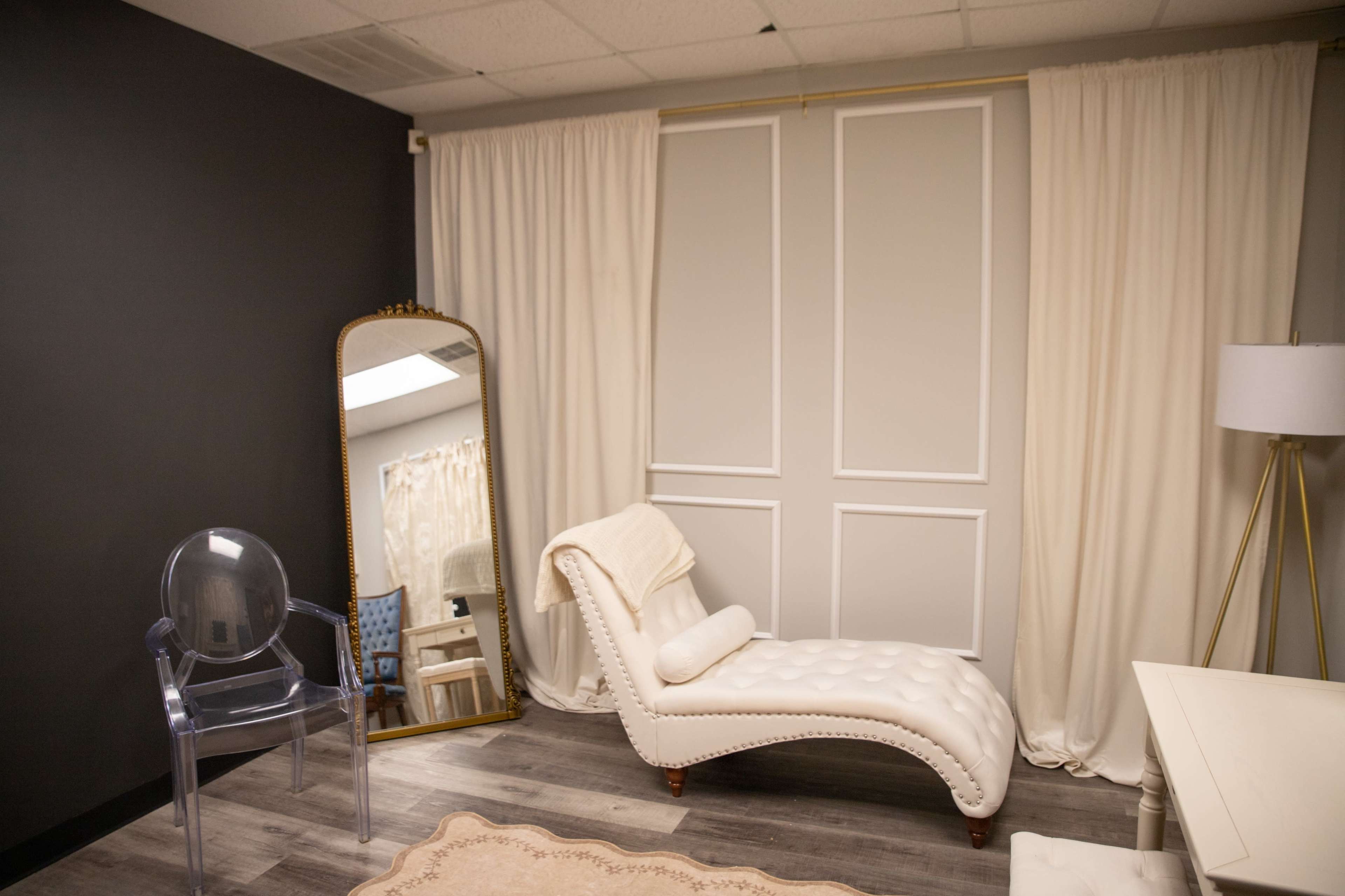 The image shows a softly lit interior room featuring a white chaise lounge, a large mirror, a clear acrylic chair, and a standing lamp beside a white table.