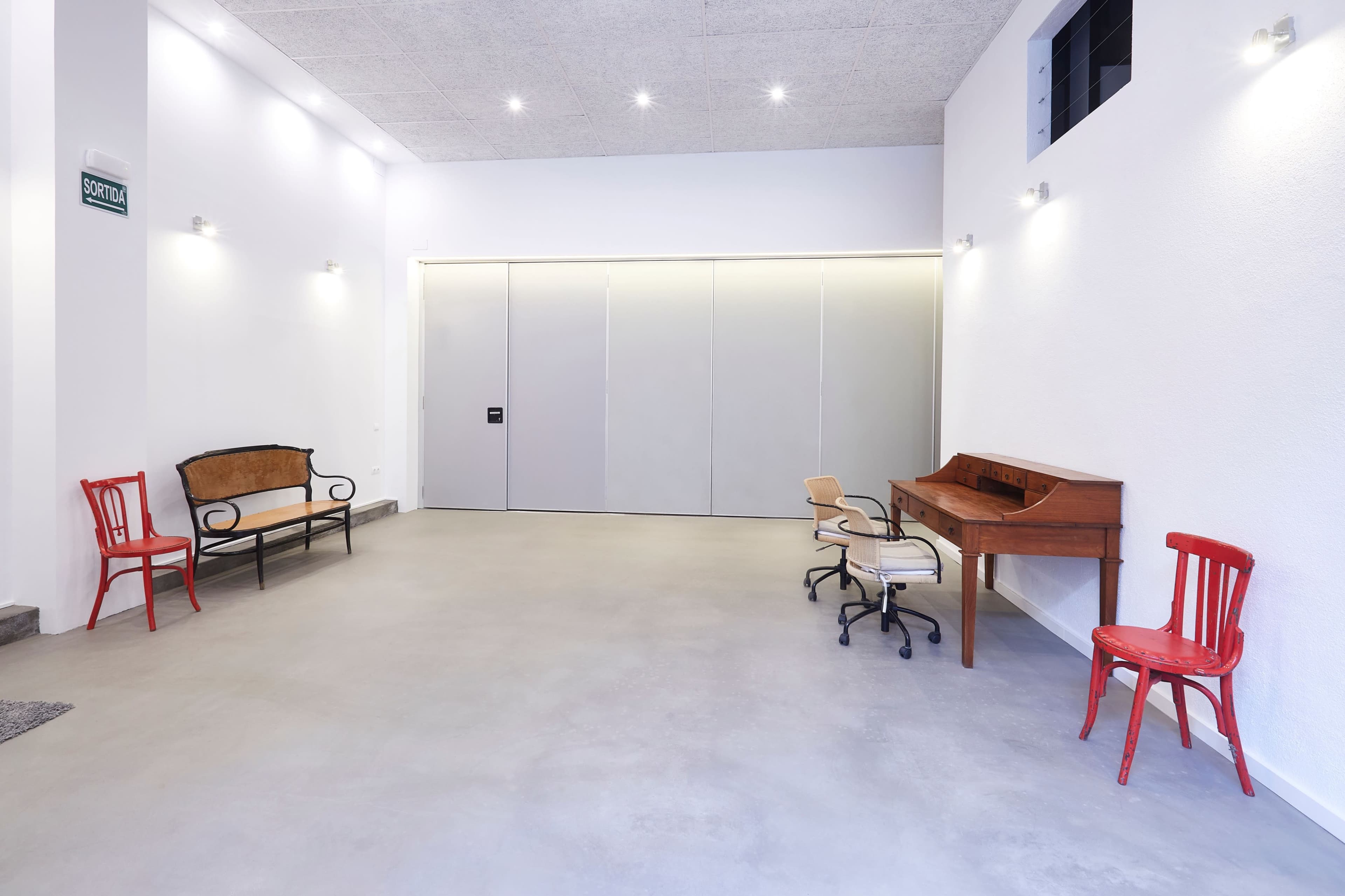 A minimalist interior space with light-colored walls, a concrete floor, a wooden desk, and several chairs, including two red ones.