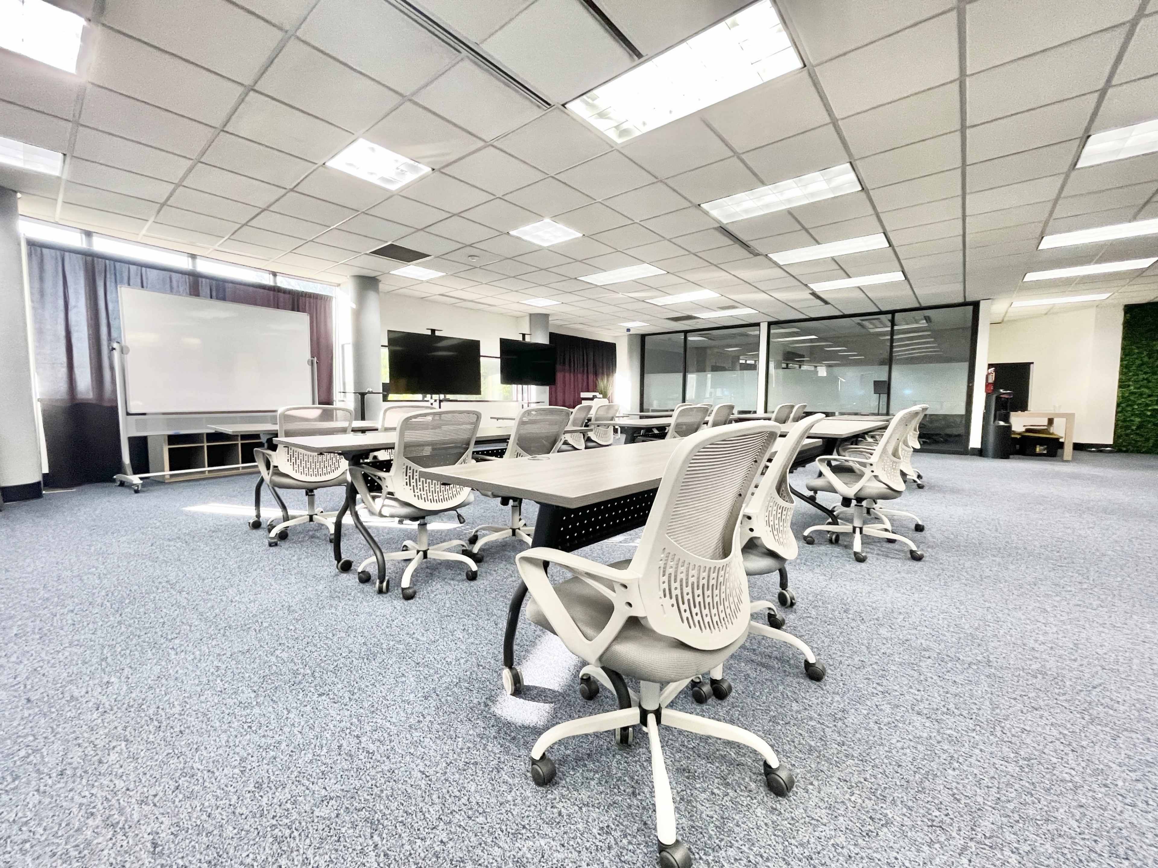 The image shows a spacious conference room with multiple tables and chairs arranged for meetings, featuring large windows and presentation screens.