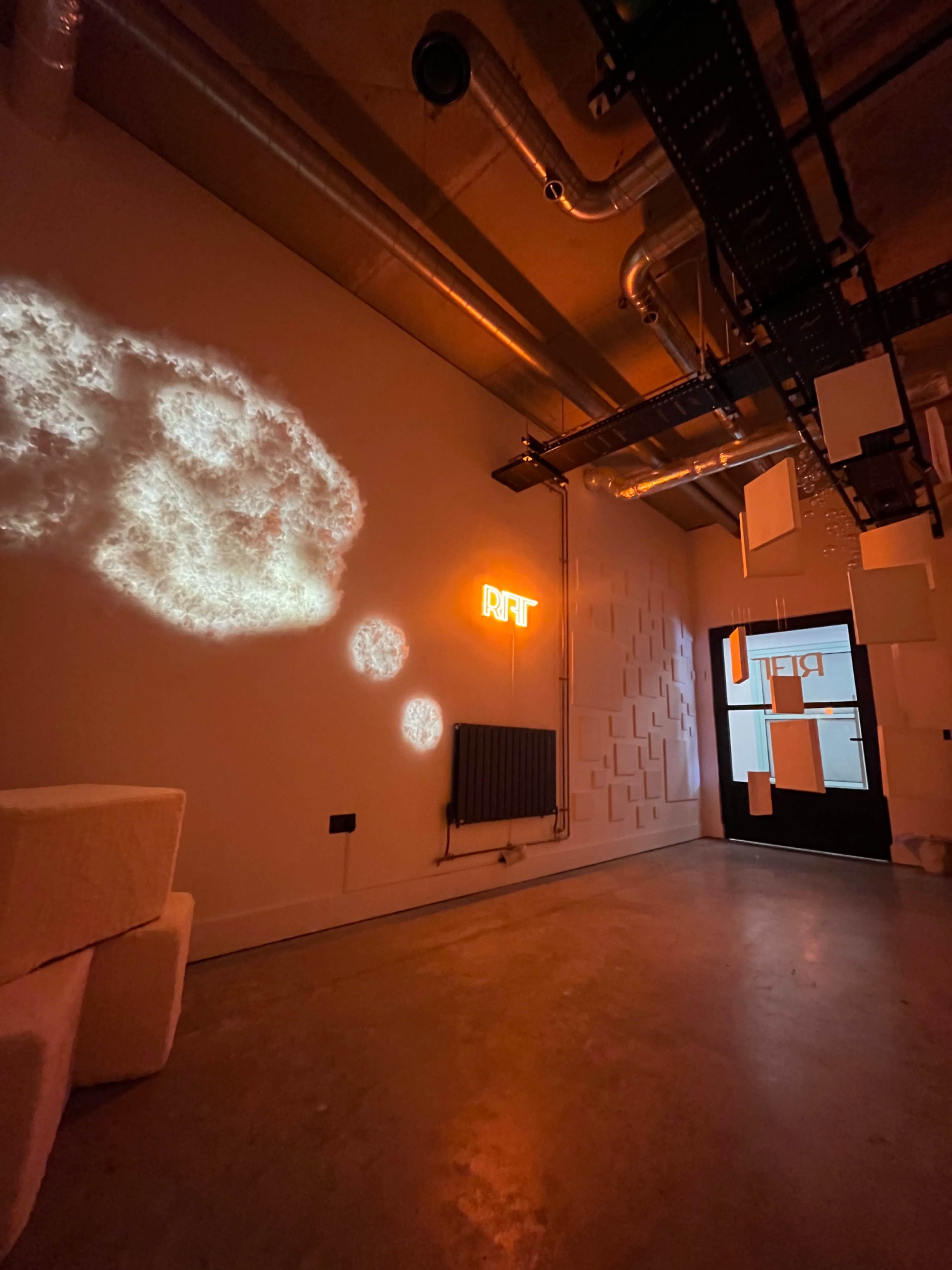 The interior of a modern room features a neon sign reading "RRT," textured wall panels, and projected visuals on the wall, with a view of a door displaying the word "PER."