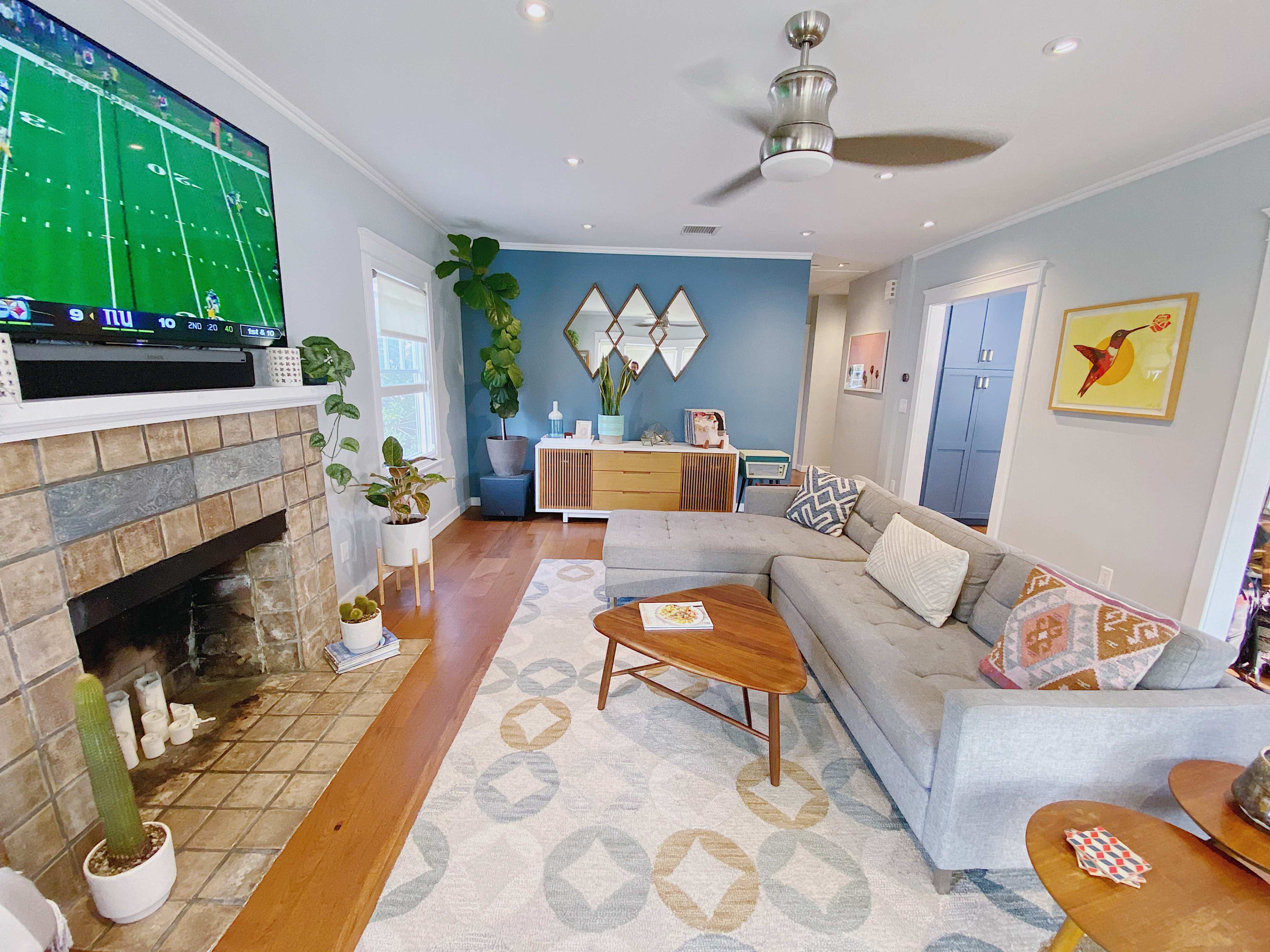 A modern living room features a gray sectional sofa, a stone fireplace, a television displaying a football game, and decorative plants.
