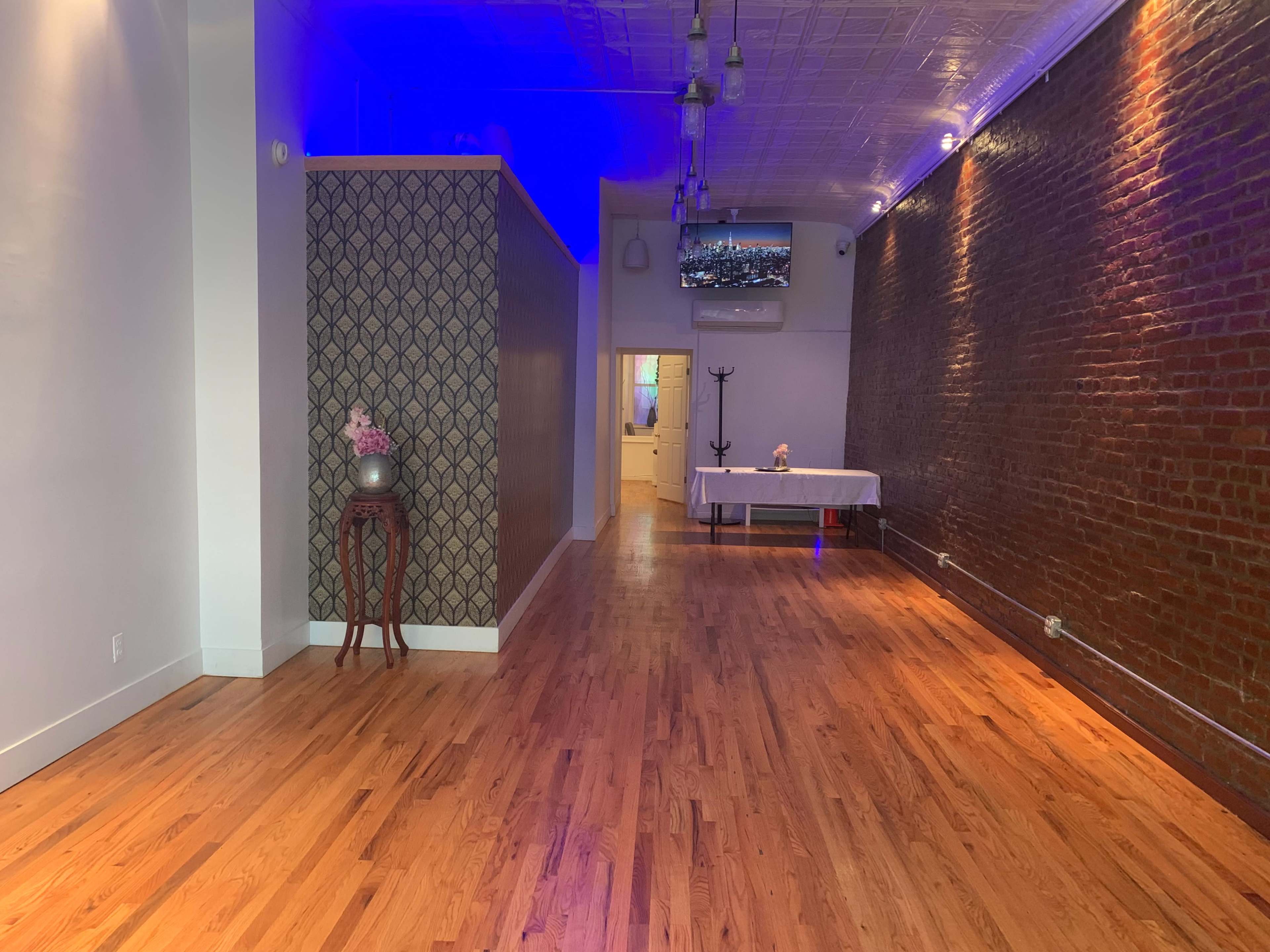 A spacious interior with a hardwood floor, exposed brick walls, and a table set up at the far end, illuminated by blue lighting.