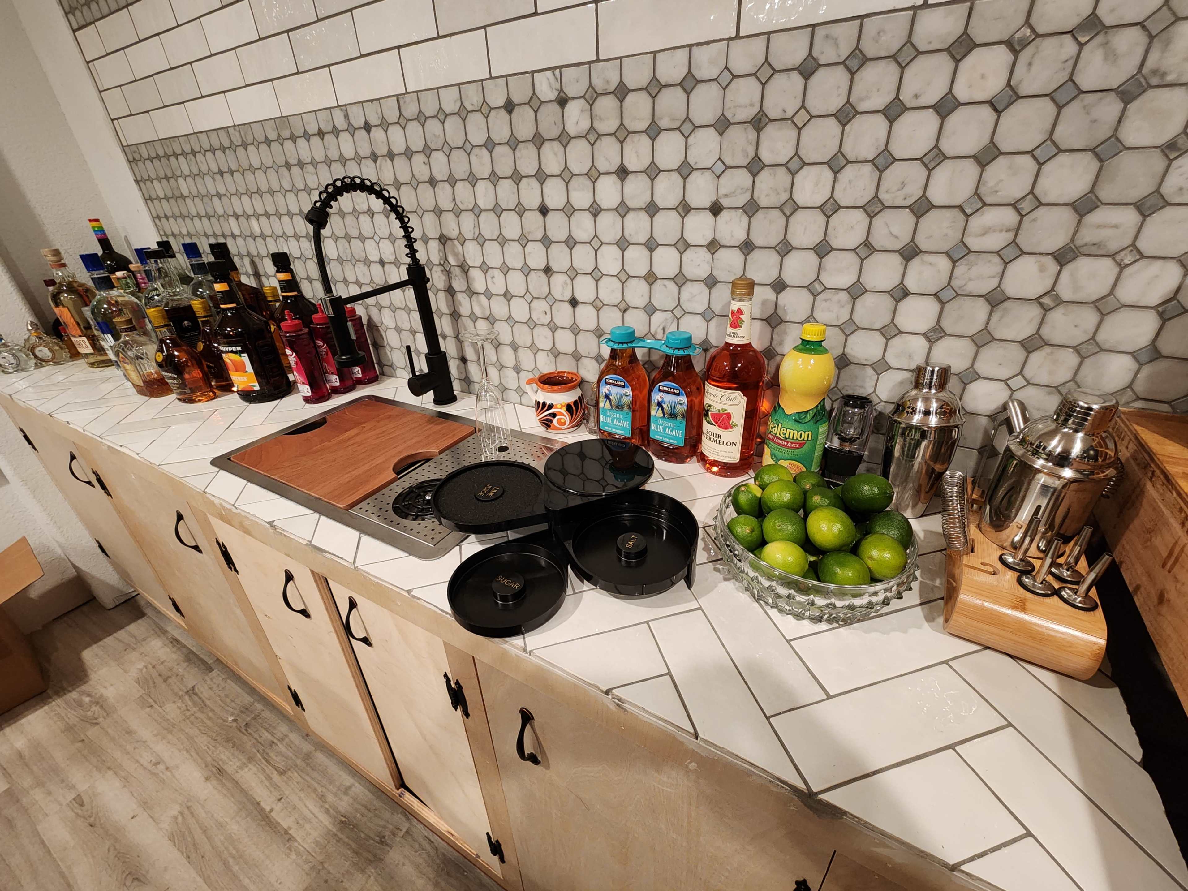 A kitchen countertop features a variety of liquor bottles, mixers, fresh limes, and bar tools arranged neatly along a tiled back wall.