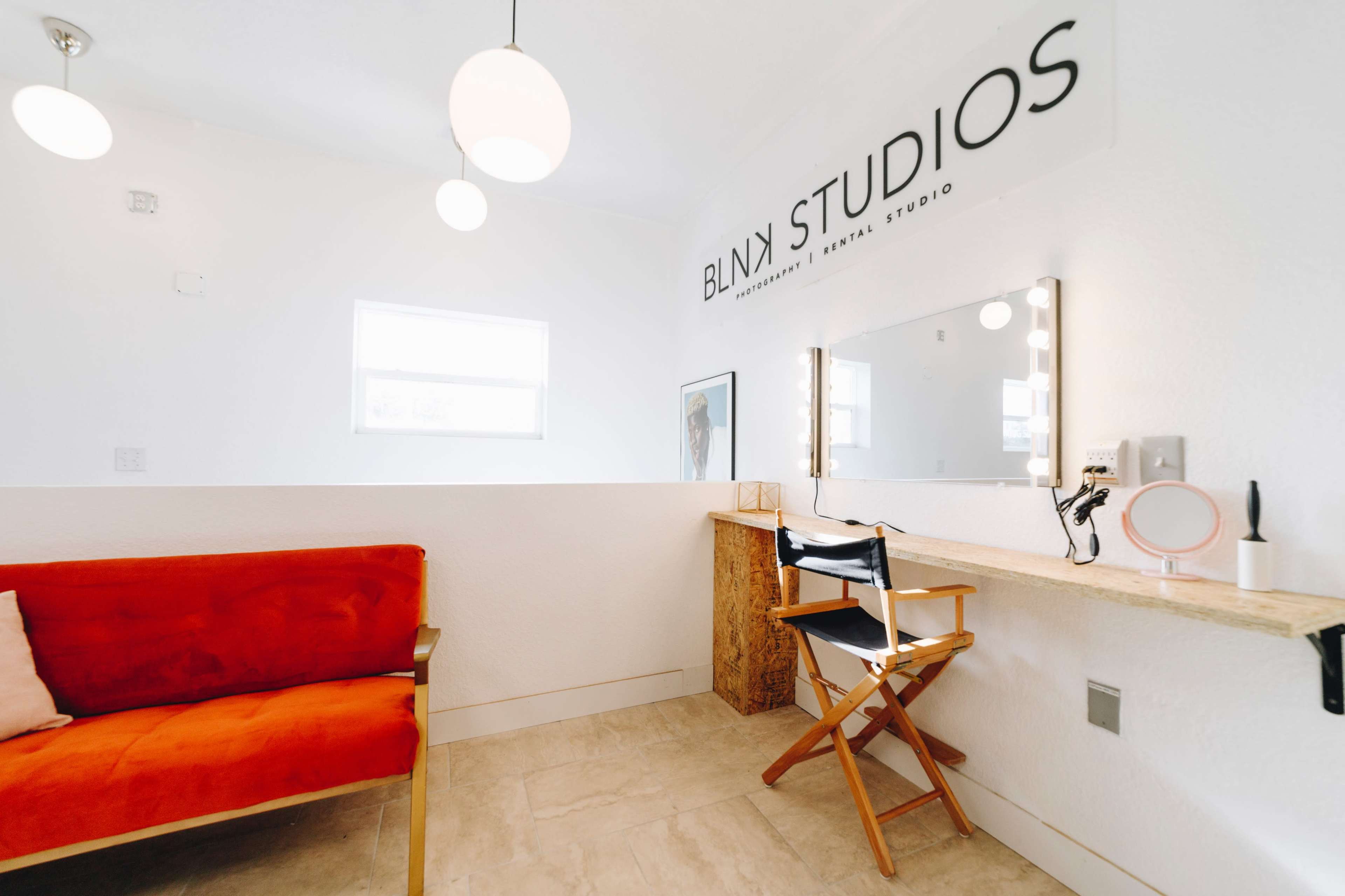 The image shows a minimalist interior of a studio space featuring a red sofa, a wooden countertop with a chair, and a mirror with lighting.