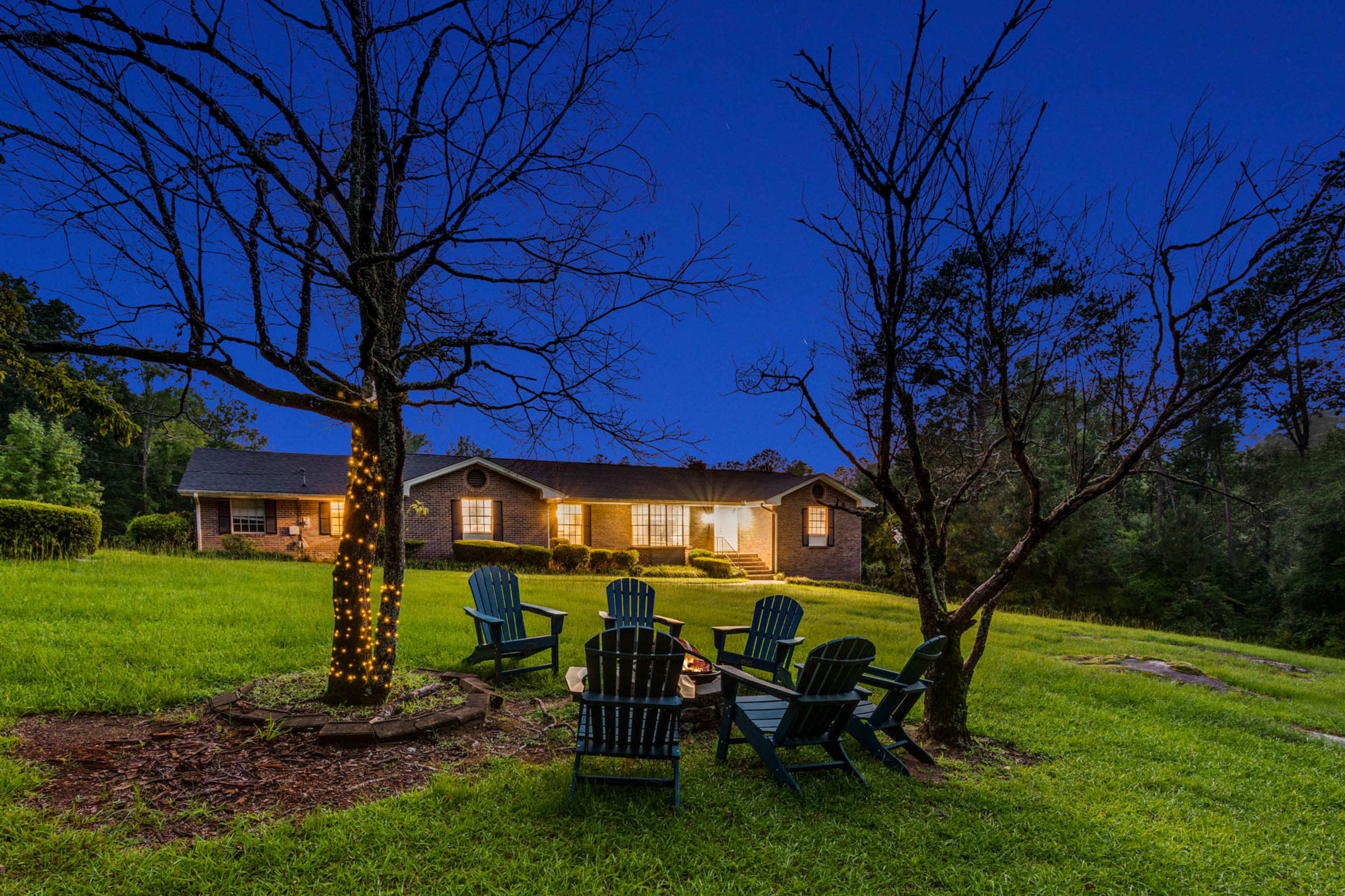 A brick house with a well-lit porch is situated on a grassy lawn, surrounded by two bare trees, with a circular arrangement of blue adirondack chairs nearby.