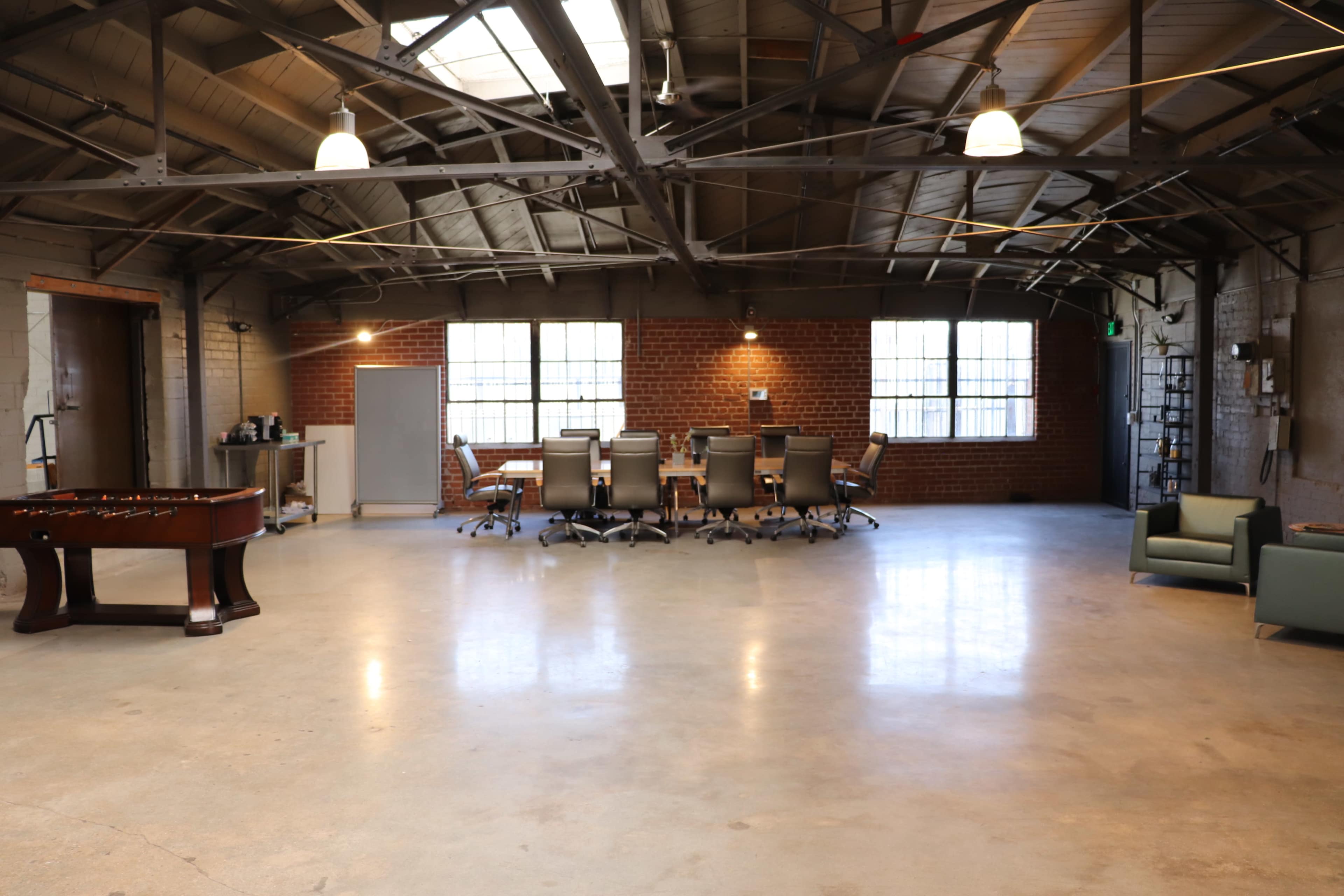 A spacious, industrial-style office features a long conference table with chairs, a foosball table, and two green sofas against the brick walls.
