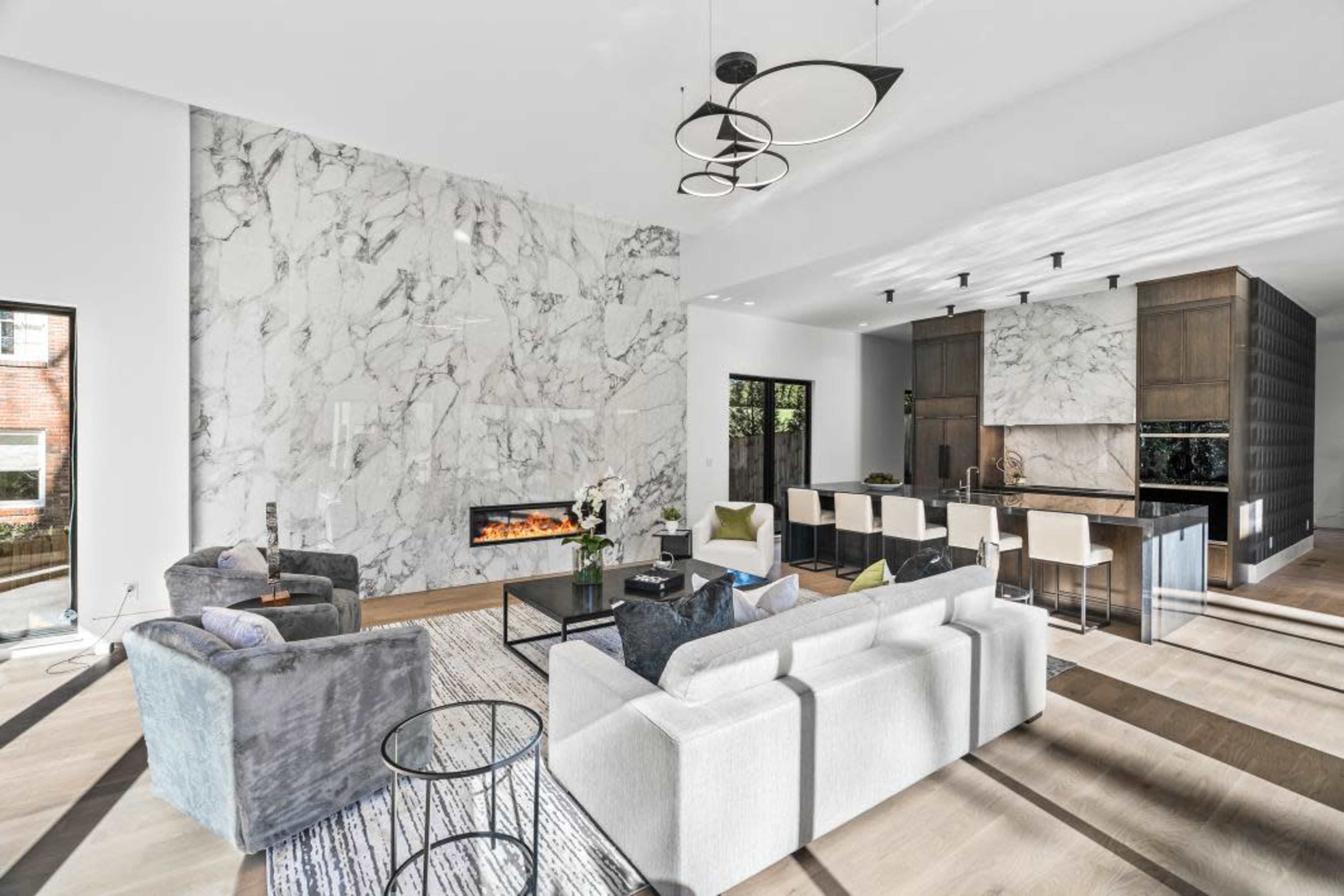 The image shows a modern living room featuring a marble accent wall, a sleek fireplace, and an open kitchen area with minimalist furniture and neutral decor.