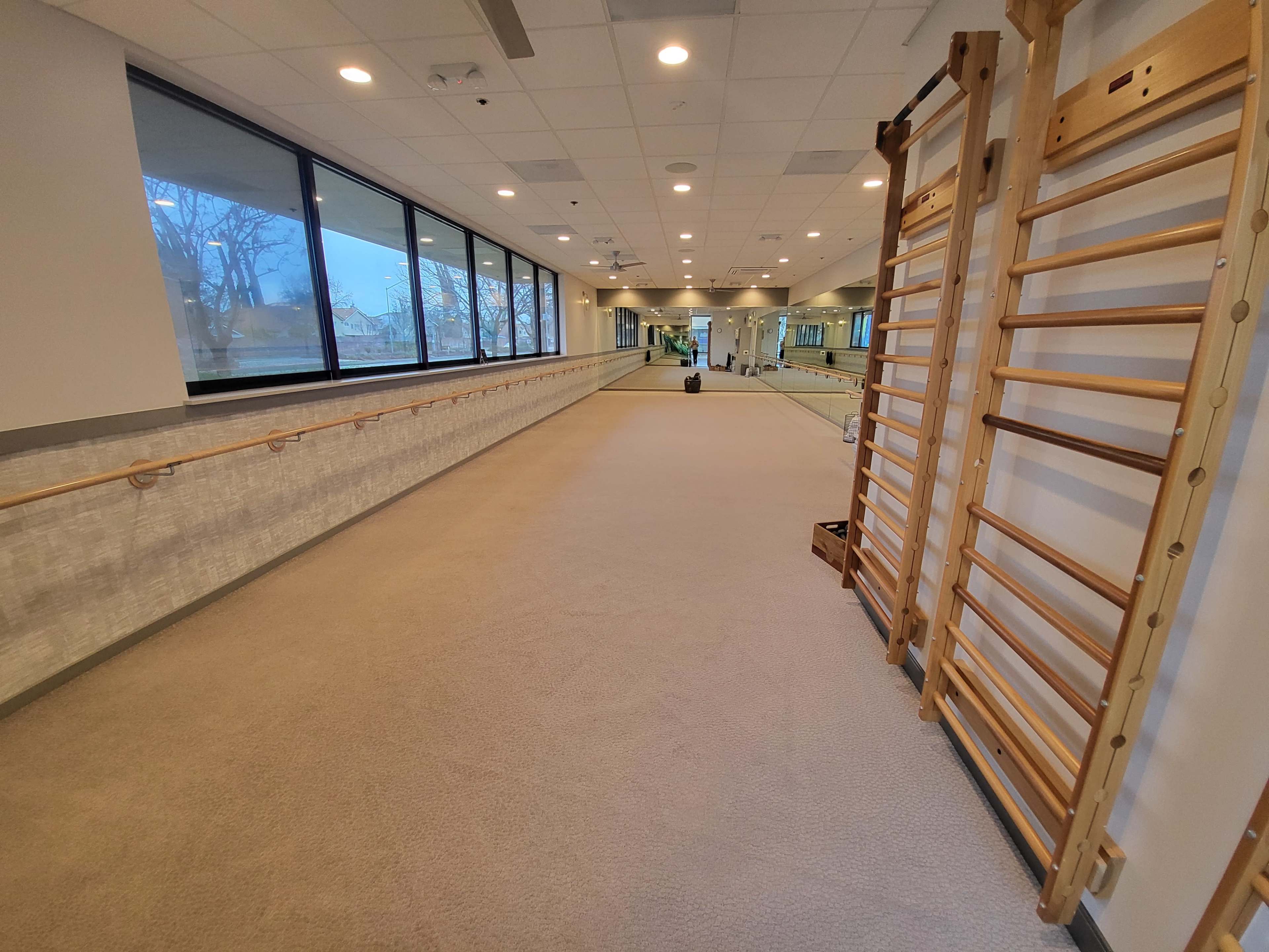 A spacious, well-lit room featuring a long mirrored wall, wooden exercise apparatus, and a smooth carpeted floor.