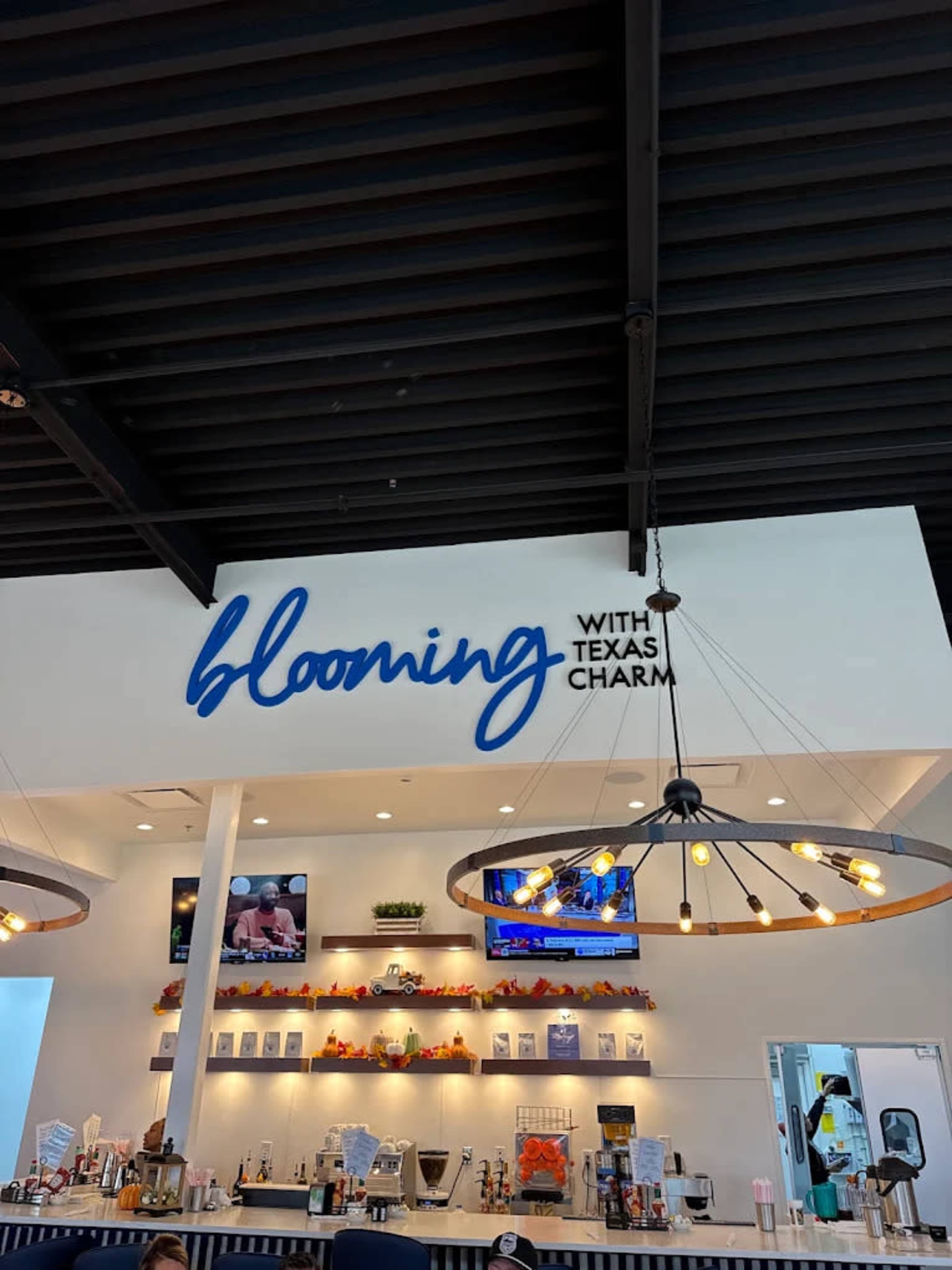 The image shows the interior of a cafe or restaurant with a prominent sign displaying the words "blooming with Texas charm" and various seating areas and decor.