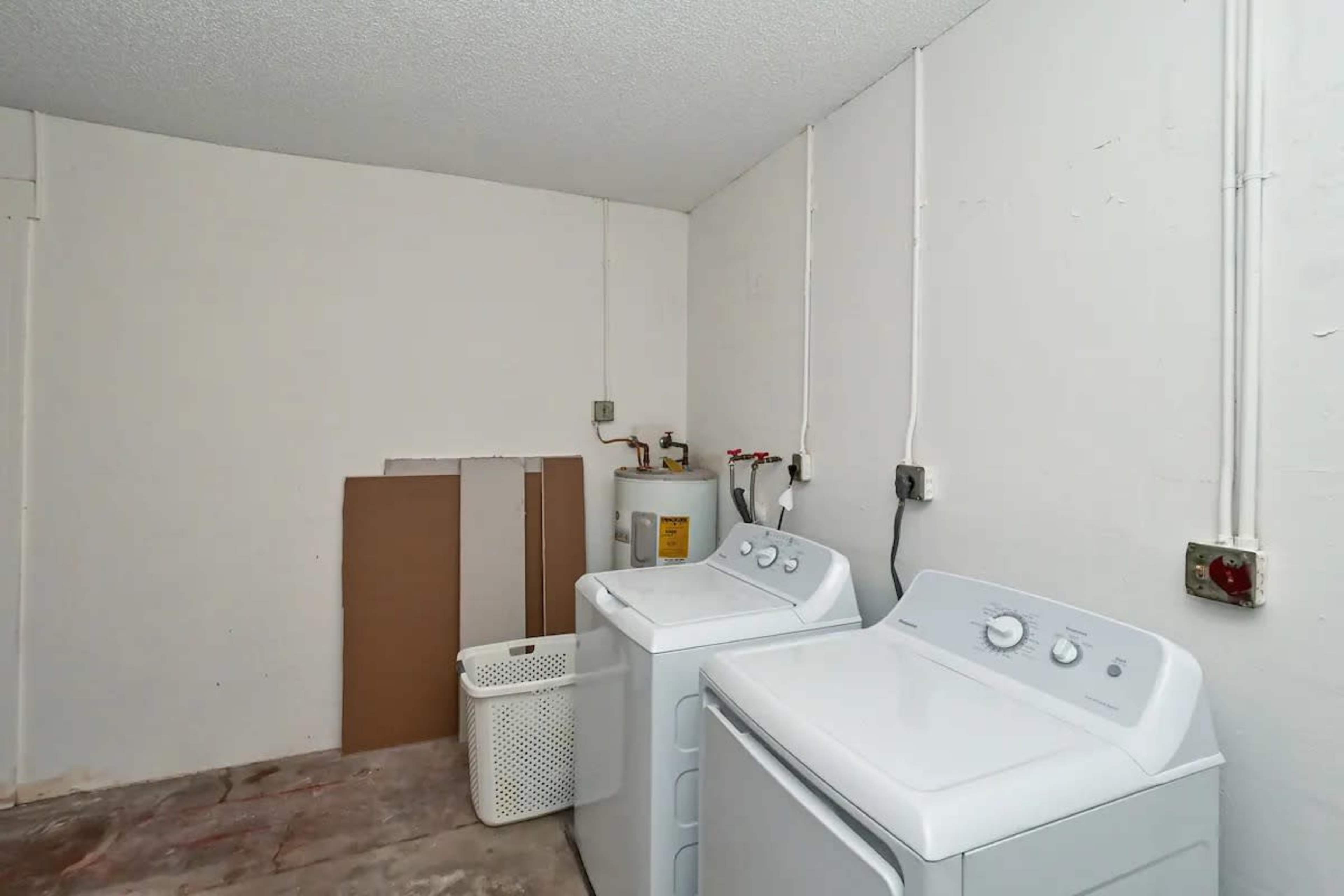 A laundry room features two white washing machines, a laundry basket, and a water heater against a plain wall.