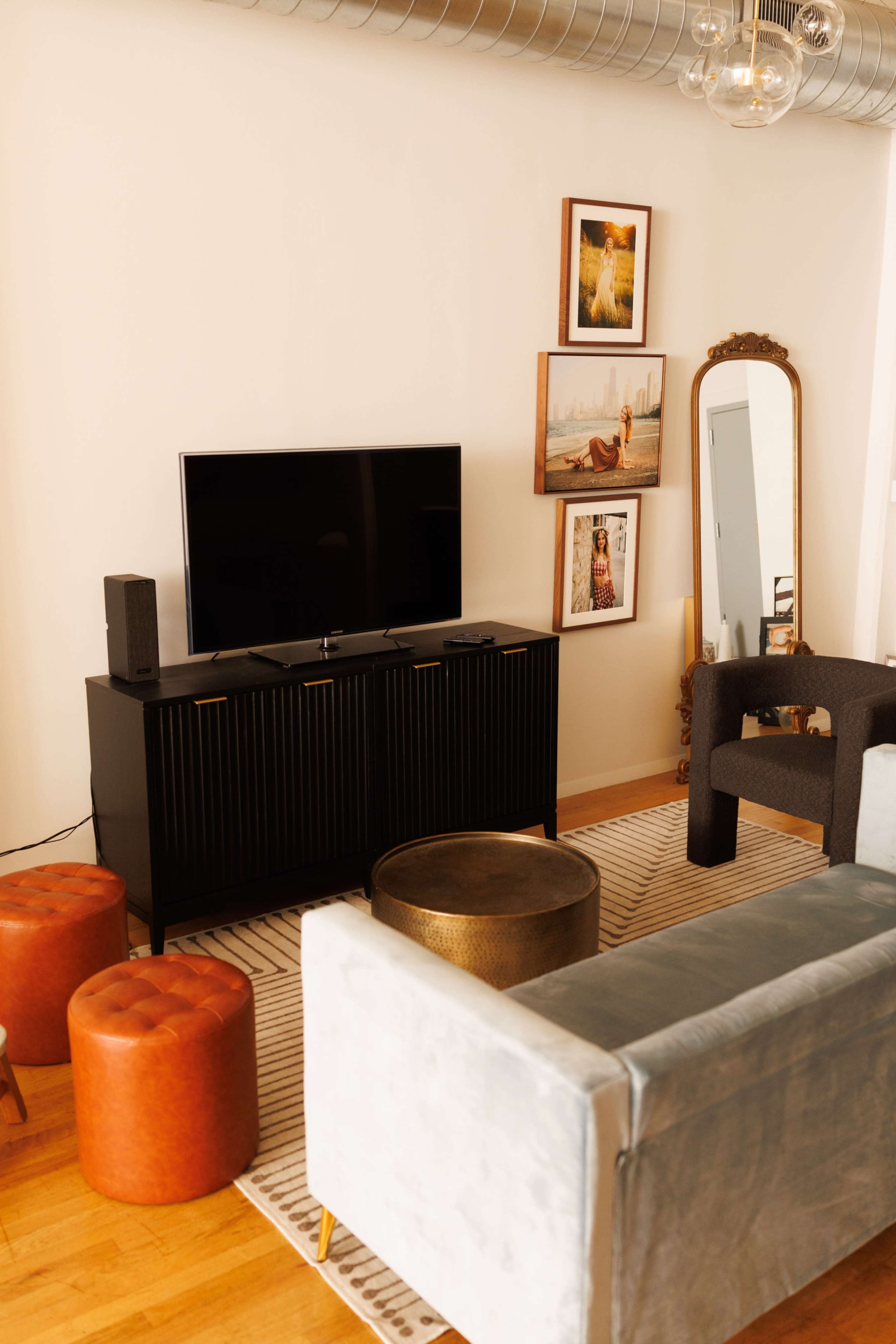 The living room features a television on a black cabinet, a large mirror, art on the wall, and various seating options including a gray sofa and round stools.
