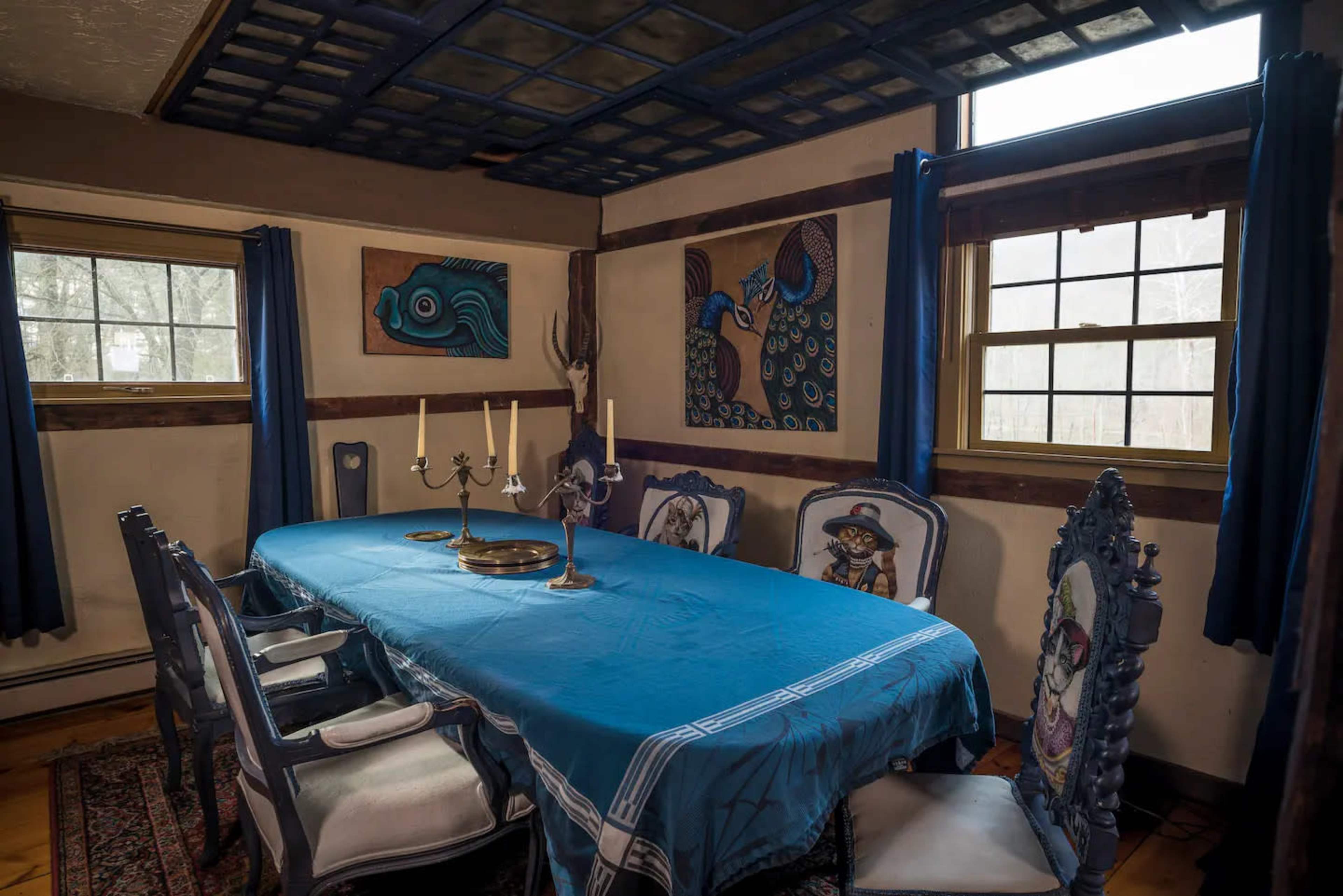 A dining room features a long blue tablecloth over a rectangular table, surrounded by ornate chairs and adorned with wall art and candles.