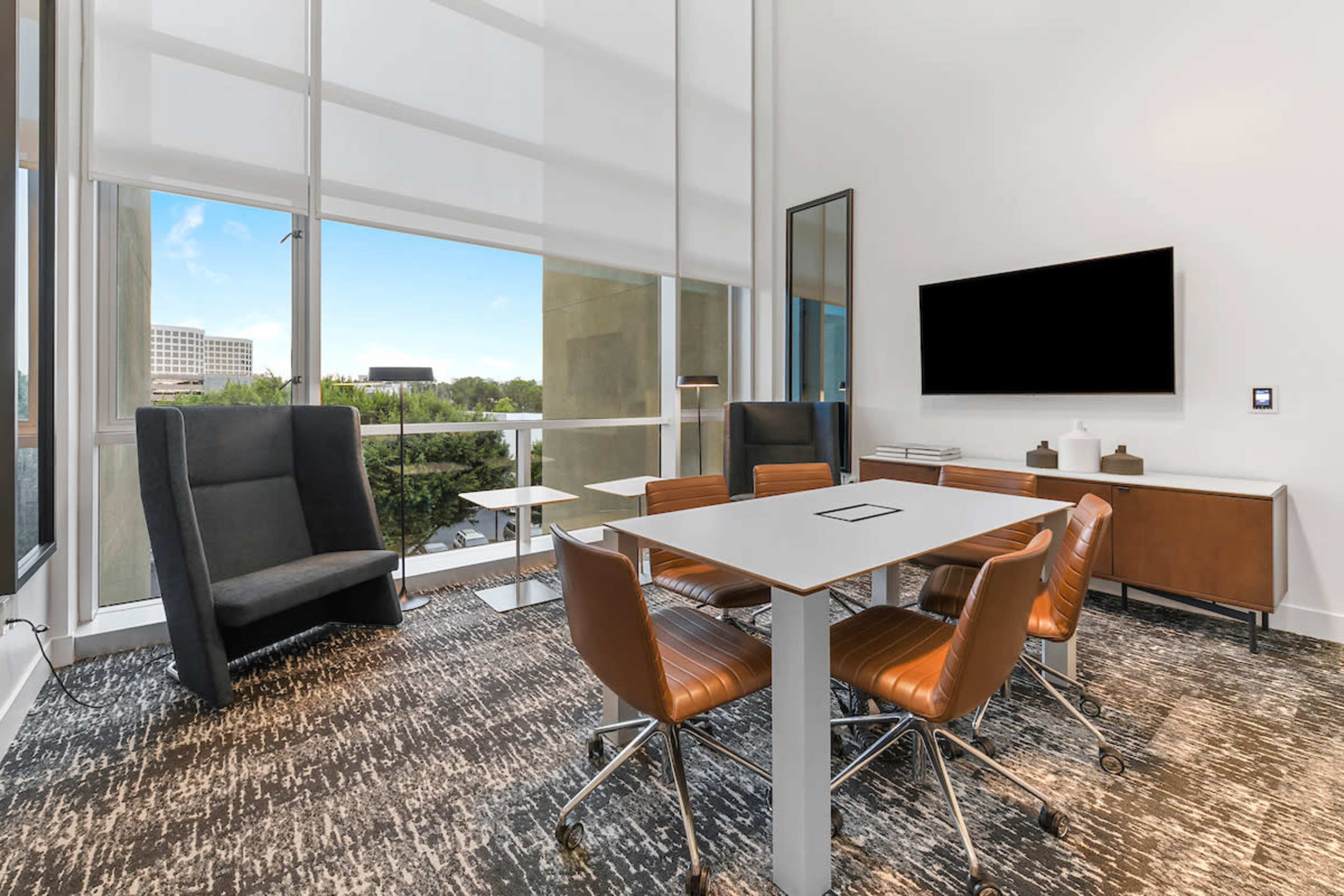 A modern conference room featuring a large table surrounded by chairs, a comfortable lounge chair, and a wall-mounted television, with large windows allowing natural light to enter.