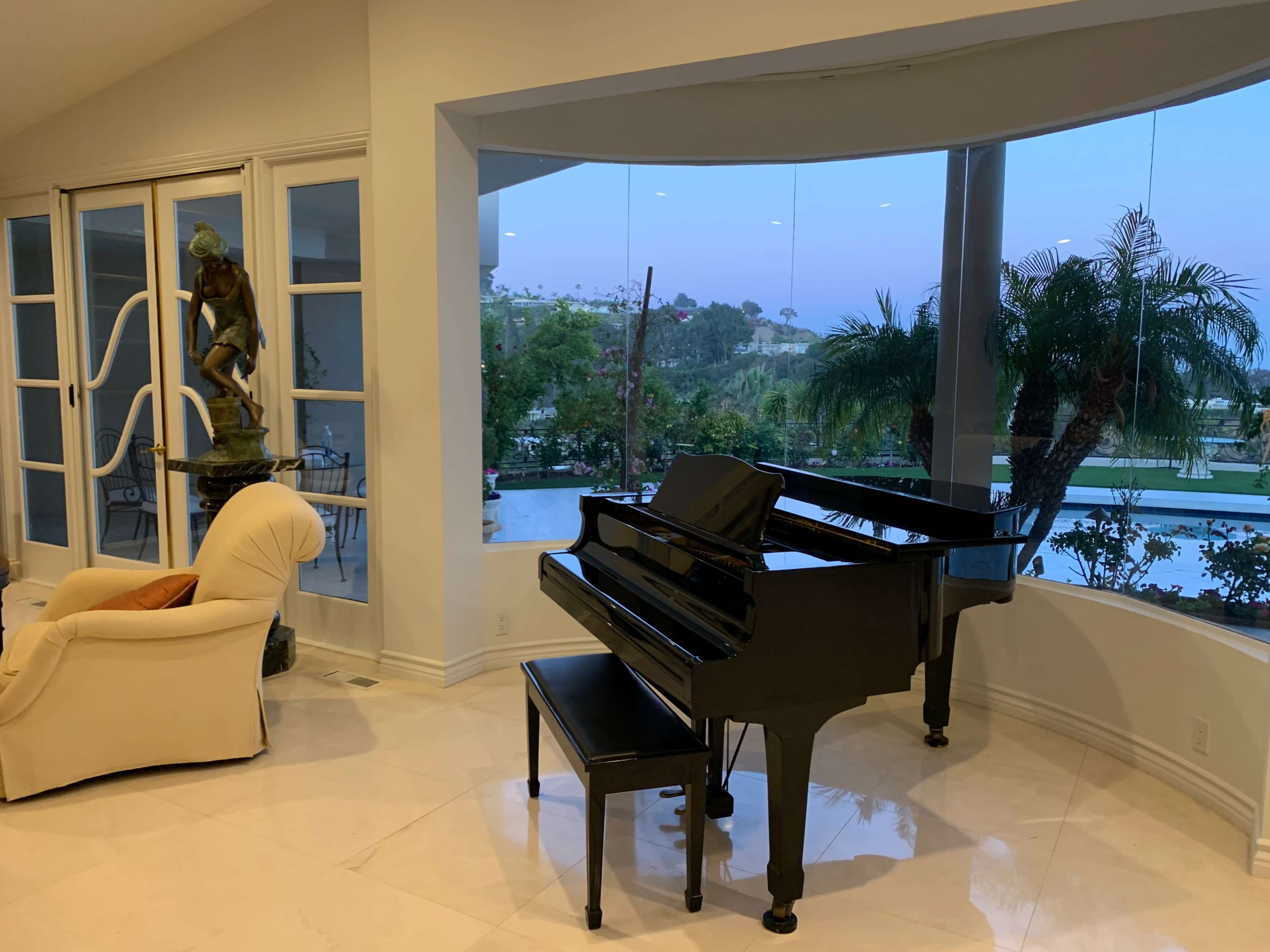 A black grand piano sits beside a white armchair in a bright room with large windows overlooking a garden and pool.