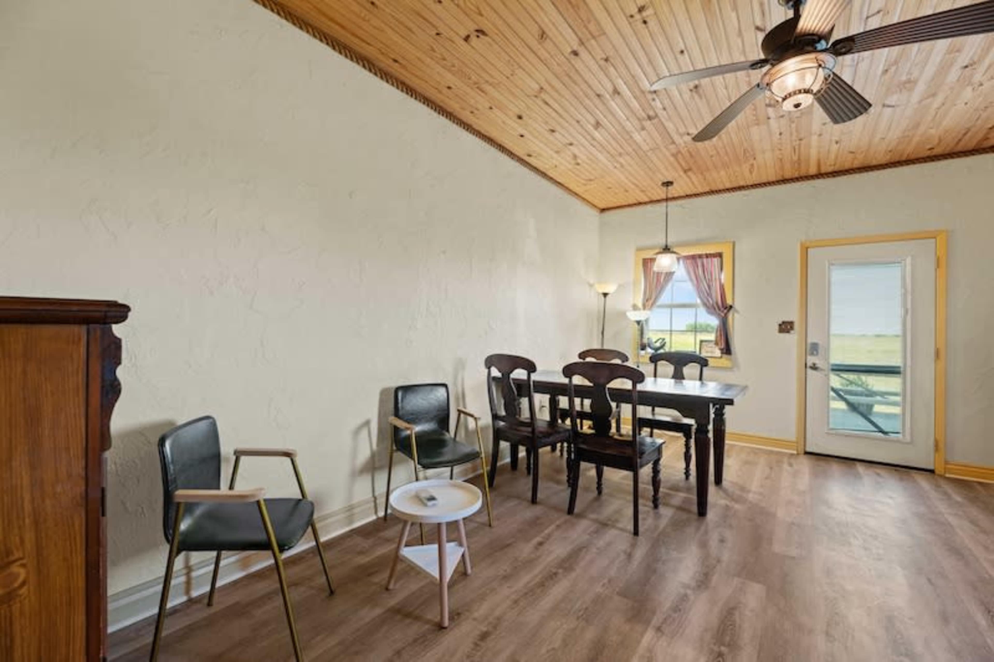 A simple dining area features a table surrounded by chairs, a door leading outside, and a window with curtains.