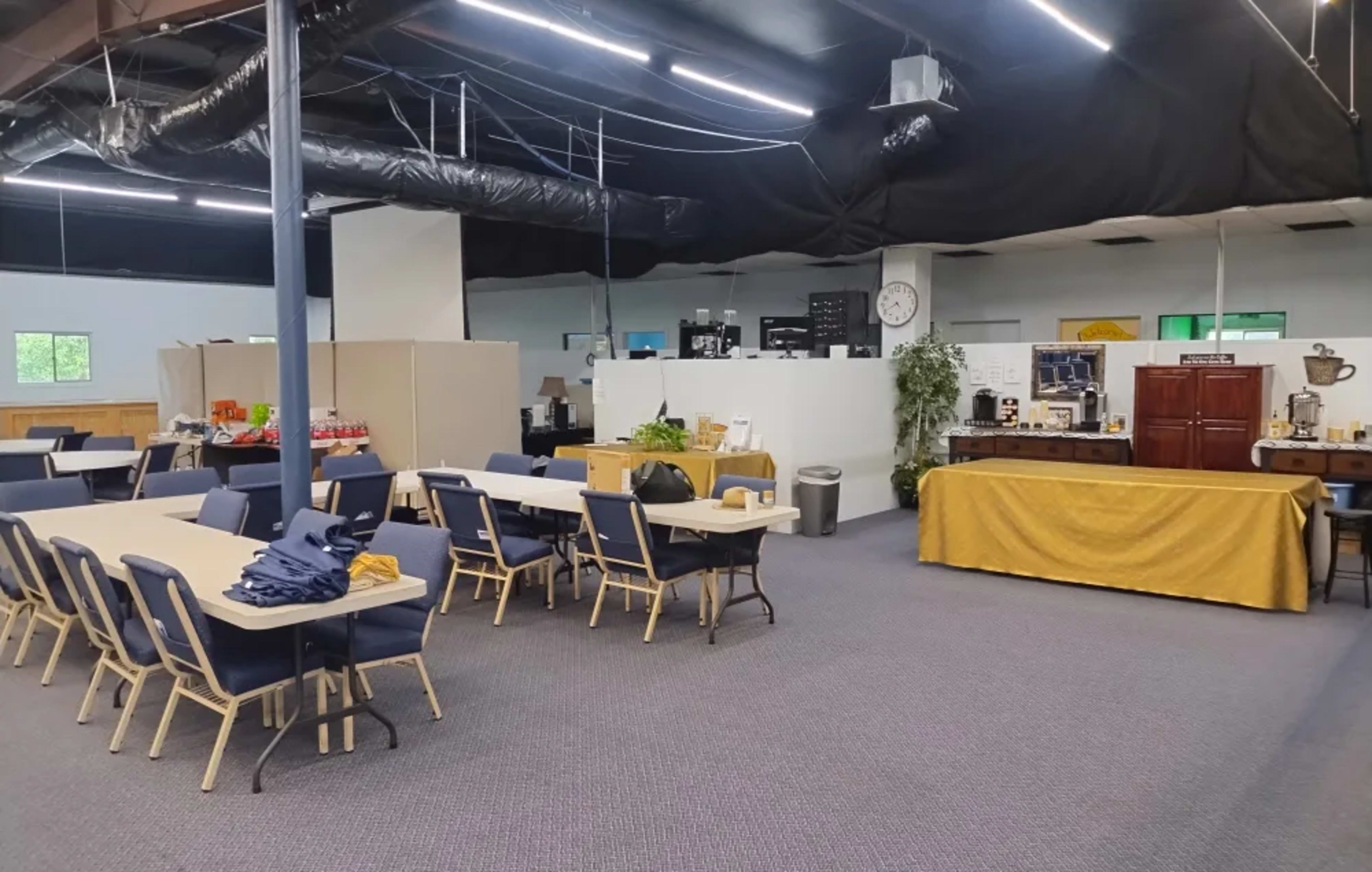 The image shows a spacious community room with tables and chairs arranged in a dining style, along with a buffet setup and various amenities in the background.