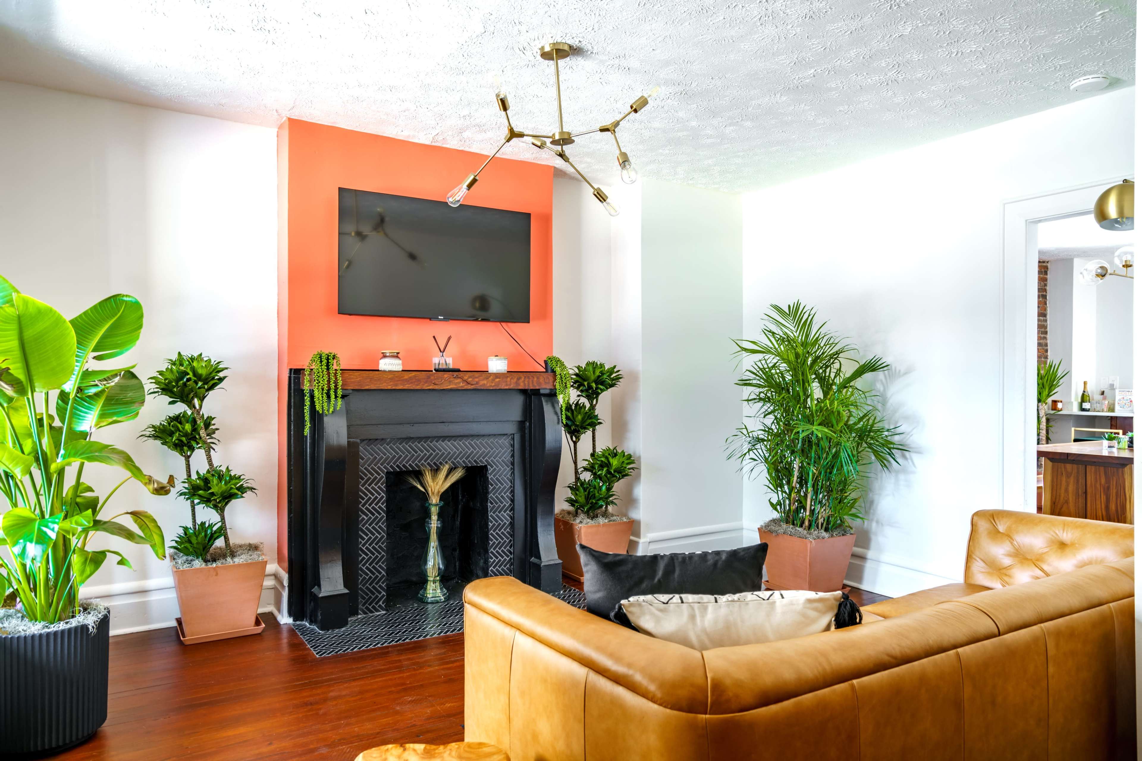 The image shows a modern living room with a tan leather sofa, vibrant orange accent wall, indoor plants, and a television mounted above a decorative fireplace.