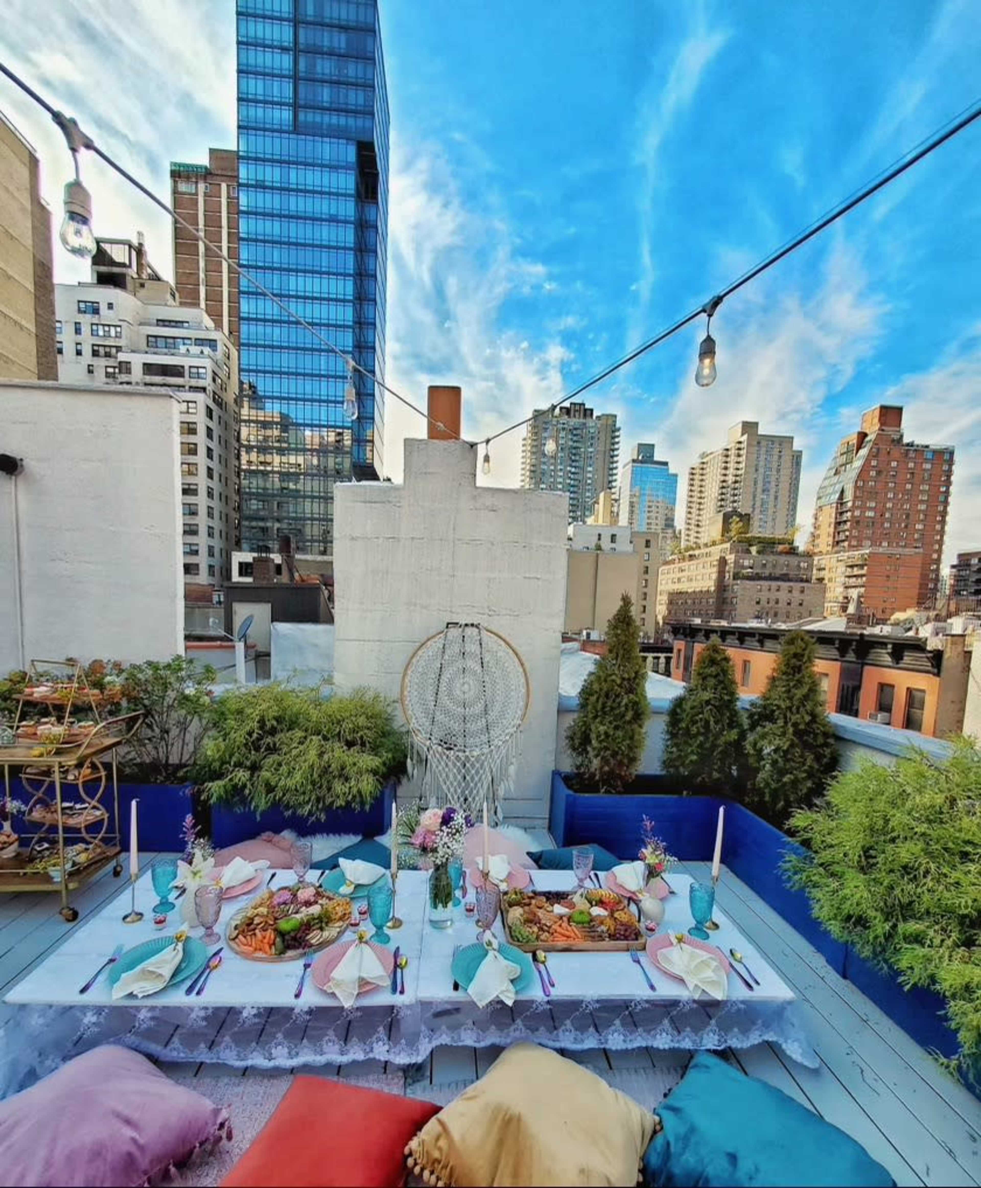 A decorated rooftop terrace features a long dining table surrounded by colorful cushions and city skyscrapers in the background.