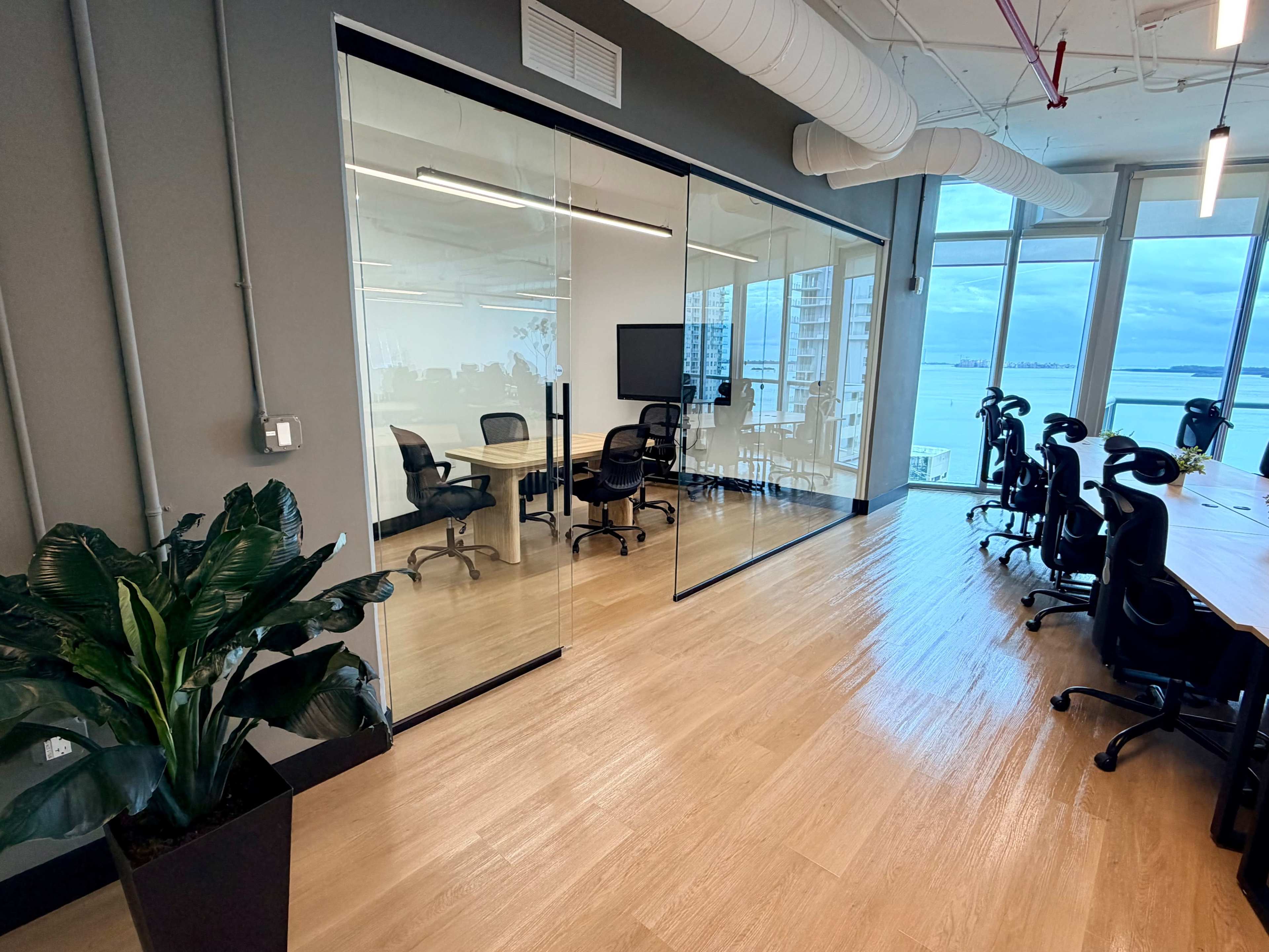 The image shows a modern office space featuring glass-walled meeting rooms, desks with ergonomic chairs, and large windows overlooking a waterfront view.