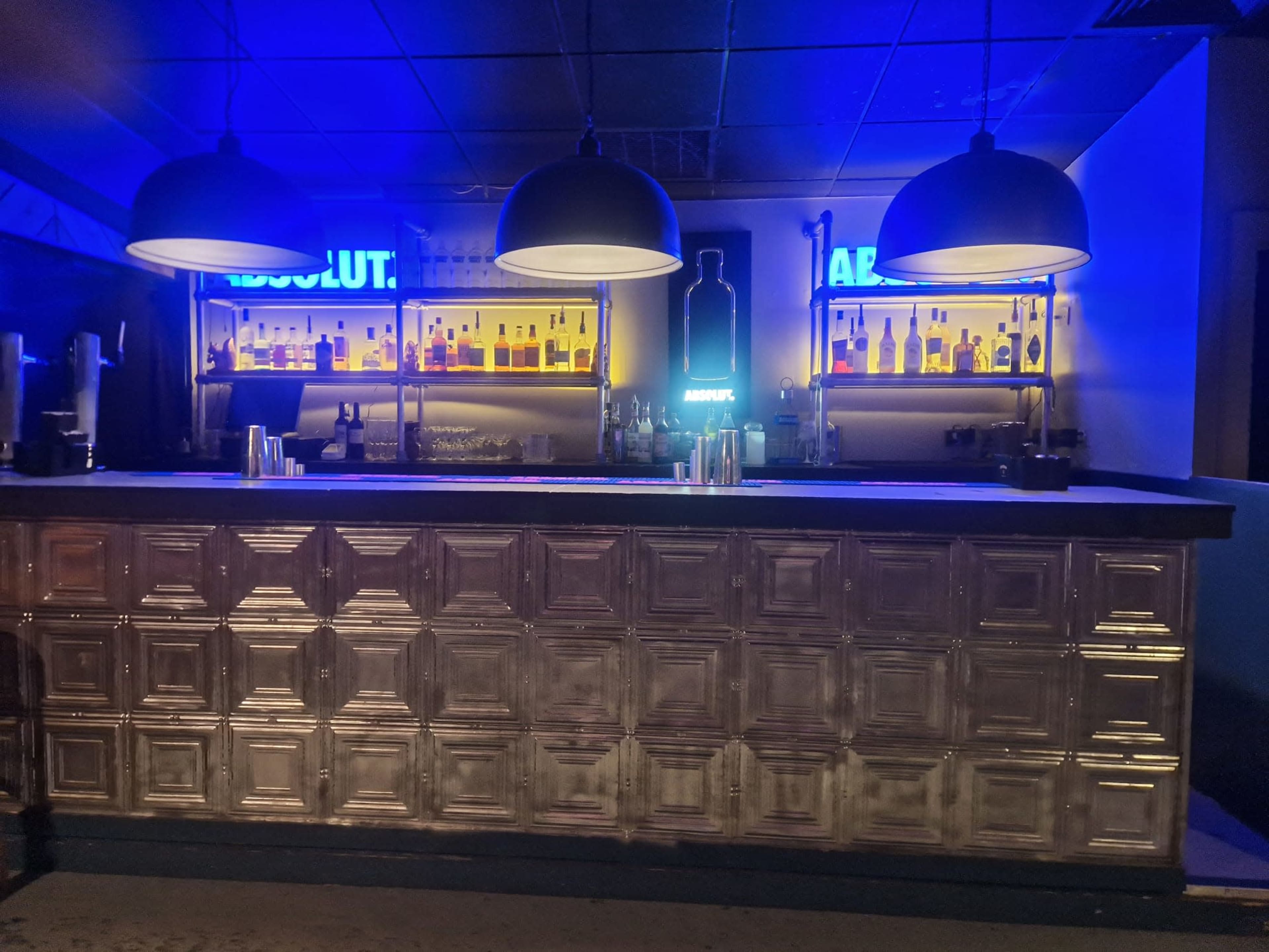 A bar with a metallic paneled front, illuminated shelves of liquor, and blue and yellow neon signs in a dimly lit setting.