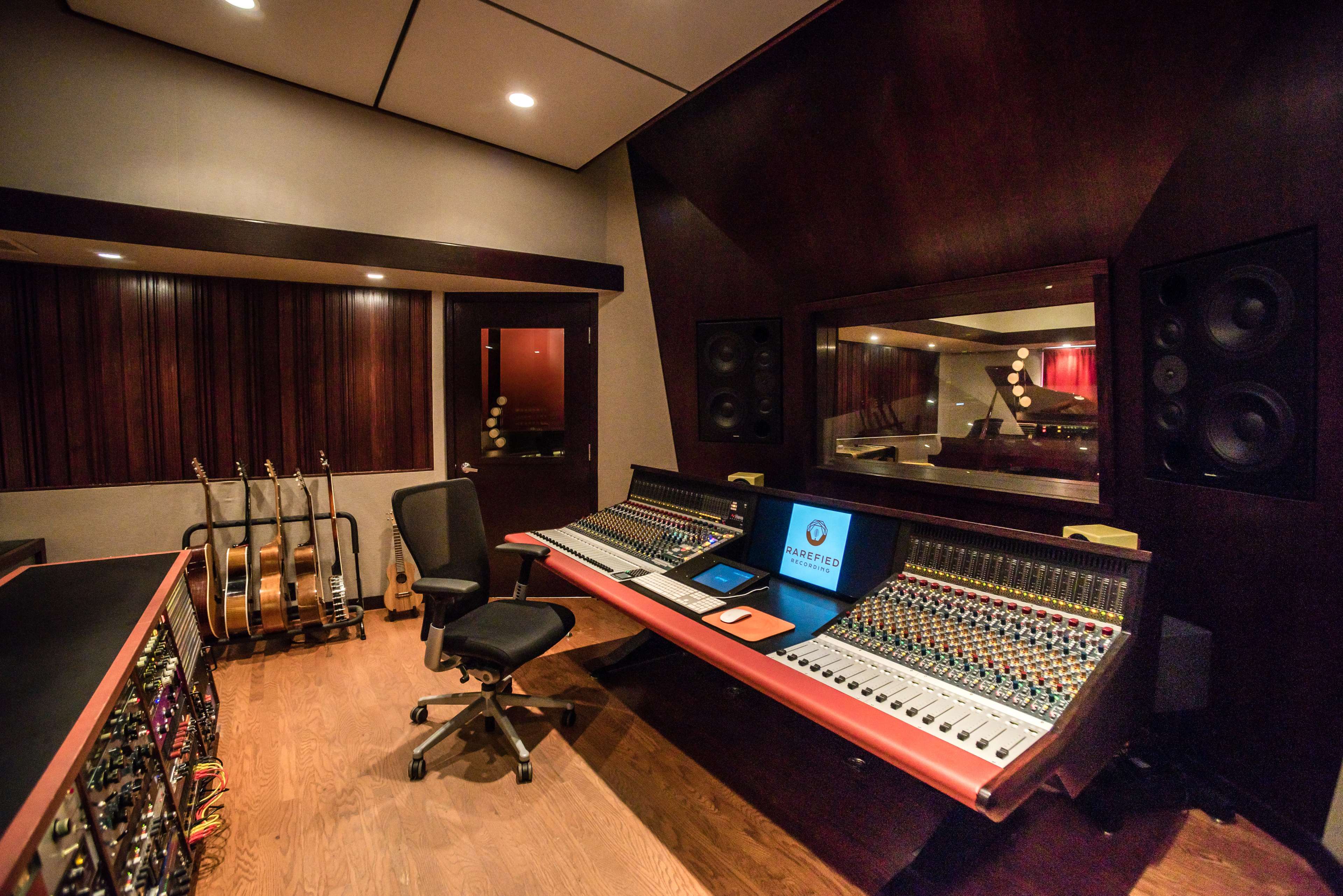 A recording studio features a large mixing console, surrounded by acoustic panels and guitars in a stand.