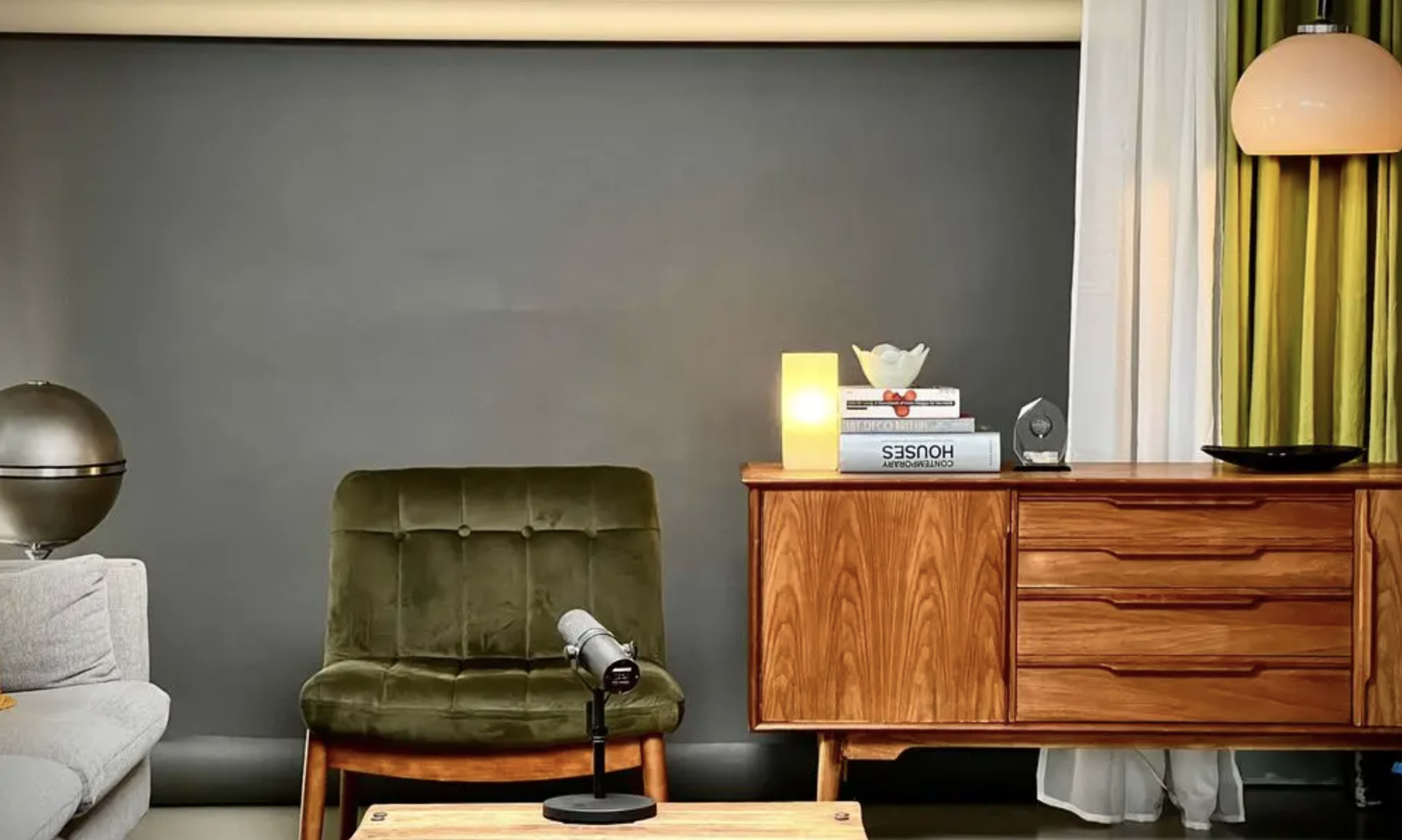 The image shows a modern living room featuring a green armchair, a wooden sideboard with books and a decorative item, and a lamp illuminating the space.