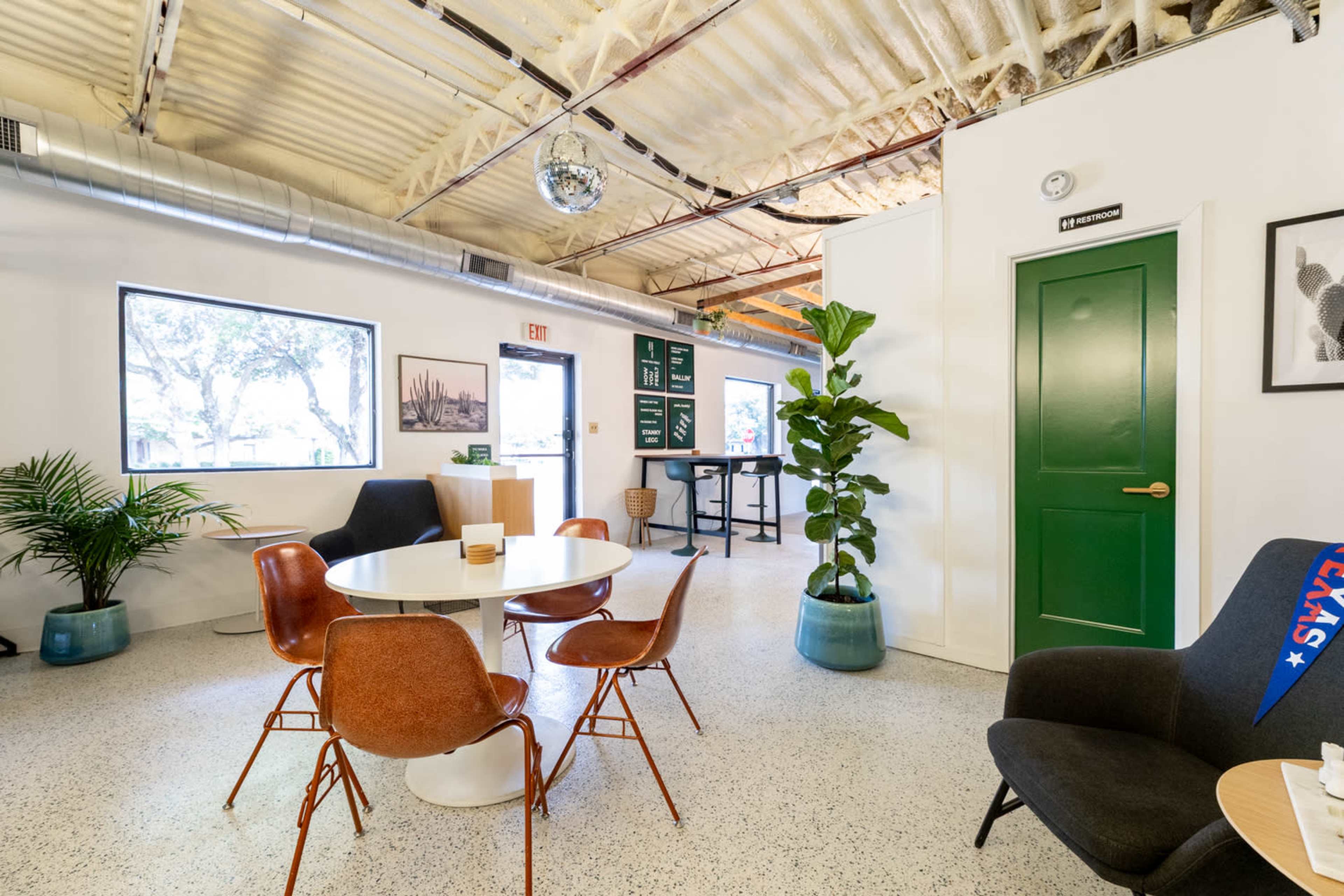 The image shows a modern, well-lit shared office space with a round table, several chairs, a plant, and a disco ball hanging from the ceiling.