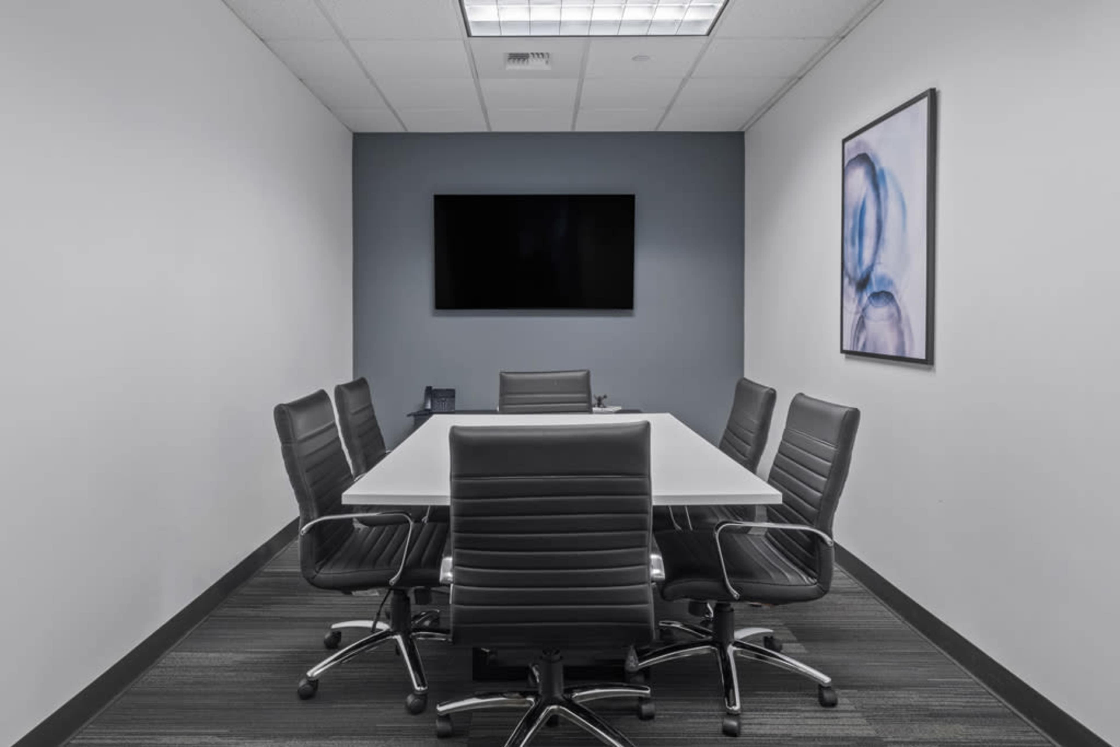 A modern conference room features a rectangular table surrounded by six black ergonomic chairs, a wall-mounted television, and an abstract art piece.