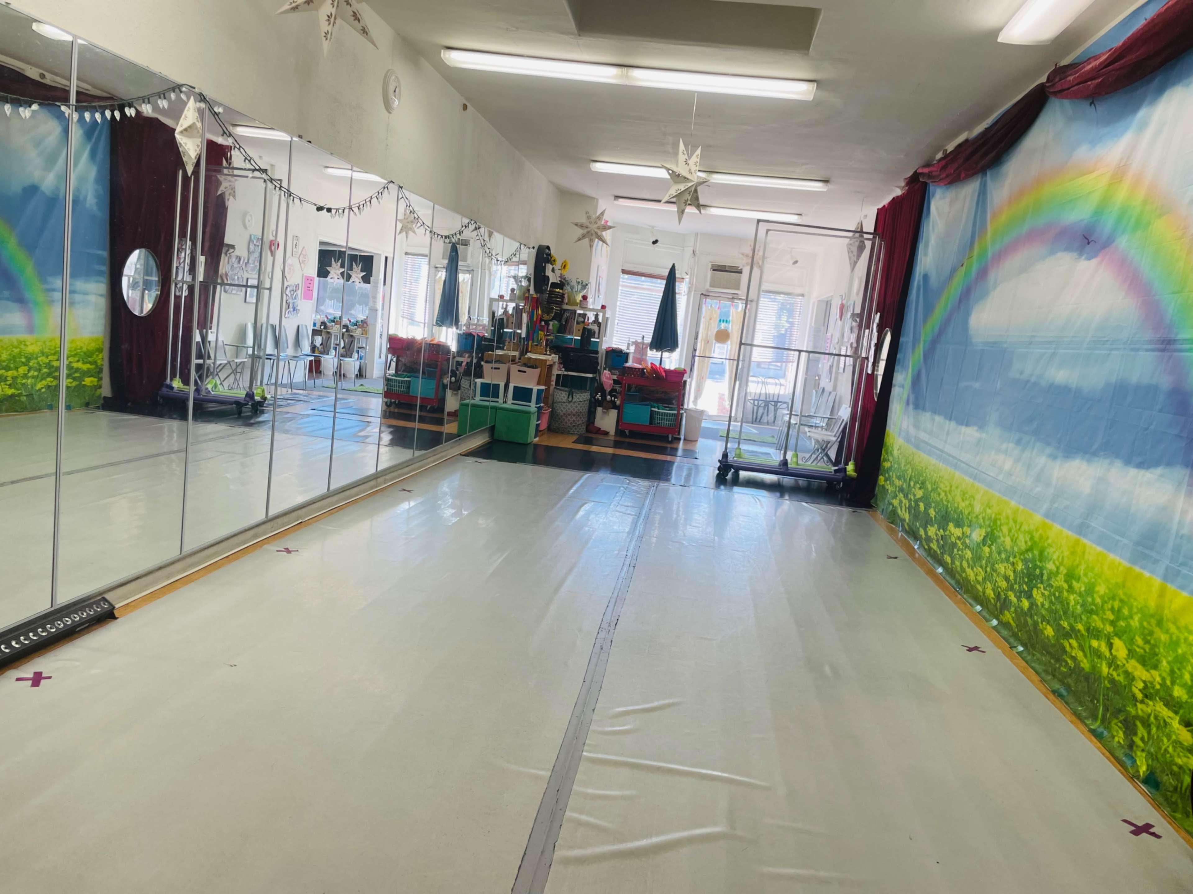 A brightly lit dance studio featuring a mirrored wall, a colorful mural of a rainbow and grass, and various instructional materials and equipment in the background.