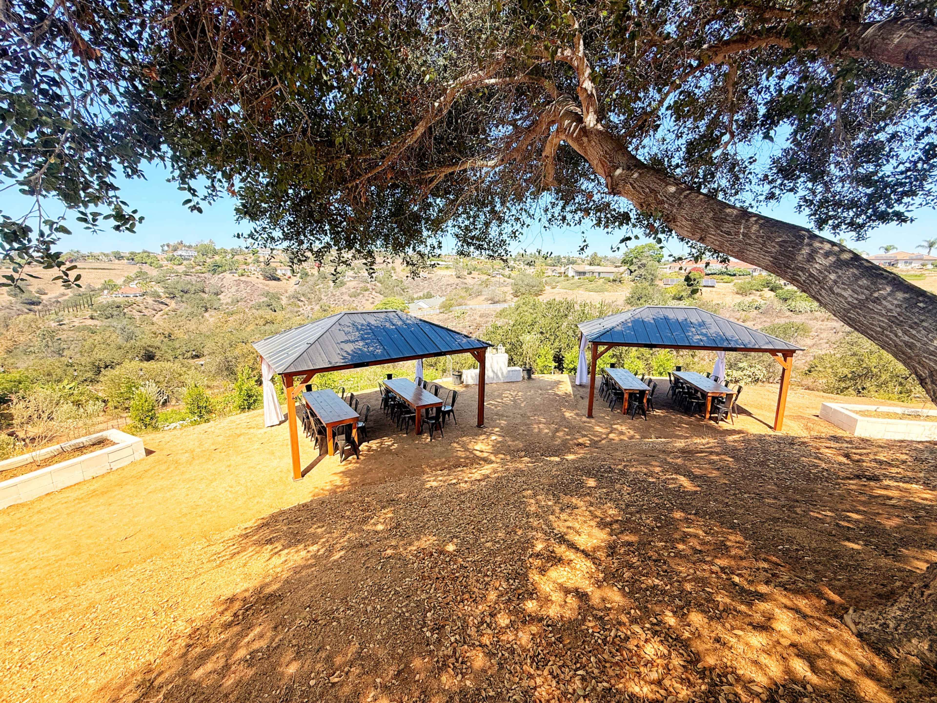 The image shows two large wooden pavilions on a dirt area overlooking a hilly landscape.