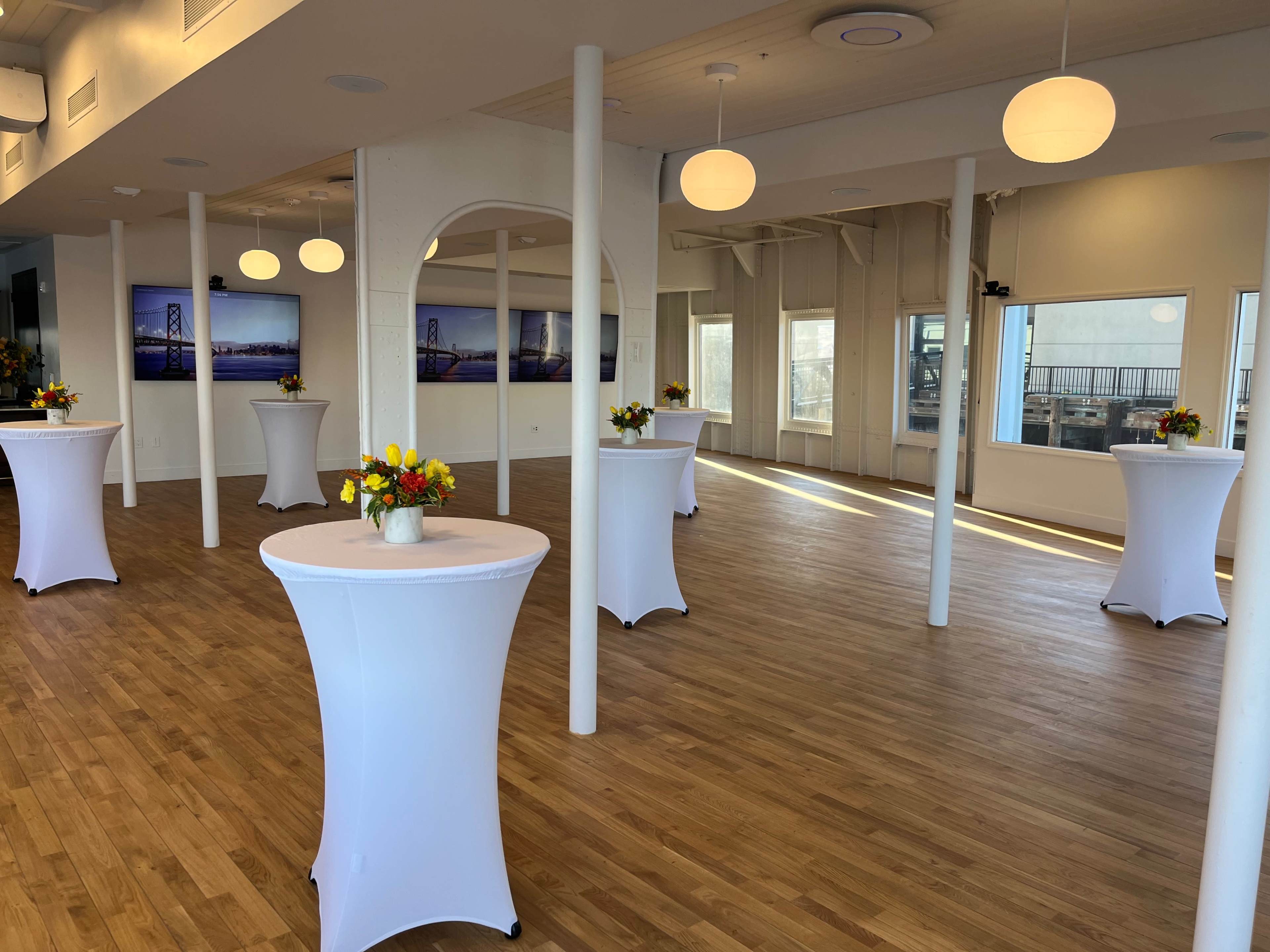 A spacious event room with several round tables covered in white tablecloths and flower centerpieces, illuminated by overhead lights and large windows.