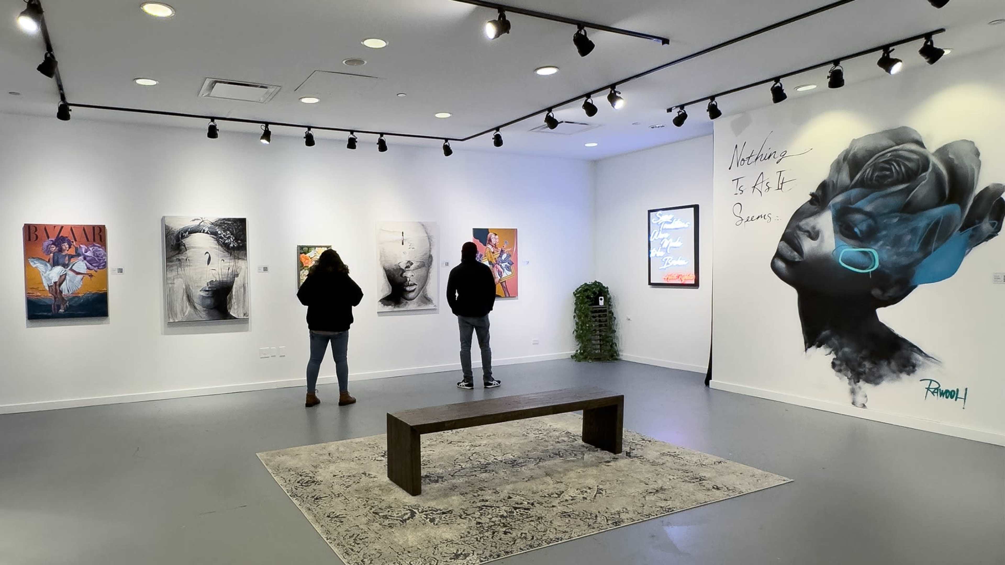 Two visitors examine artworks in a contemporary gallery featuring various framed pieces and a mural on the wall.