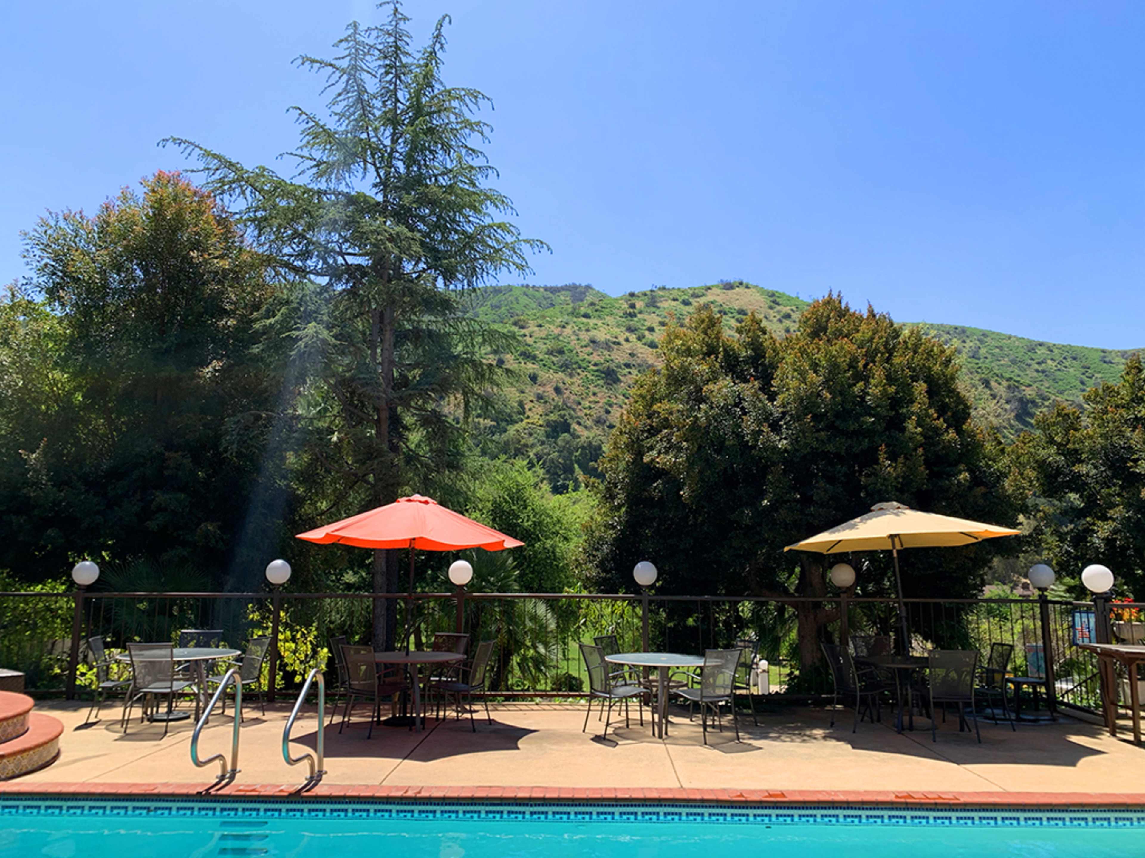 The image shows a pool area with two umbrellas and tables set against a backdrop of green hills and clear blue skies.