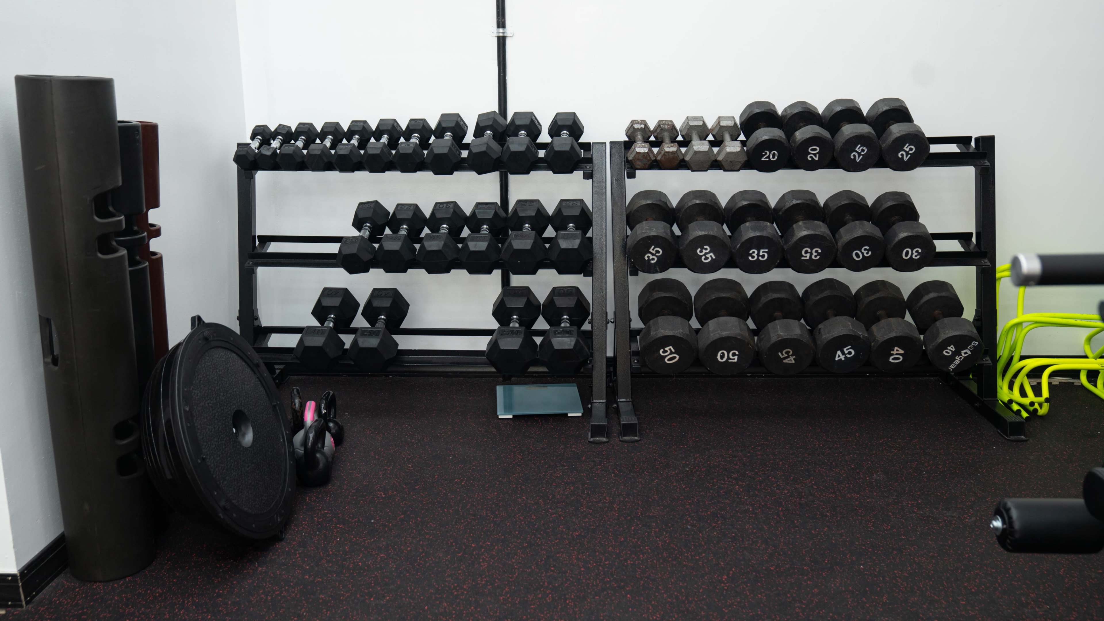 A gym area featuring two rows of neatly arranged dumbbells on racks, along with a weight plate, a kettlebell, and a scale on a rubberized floor.