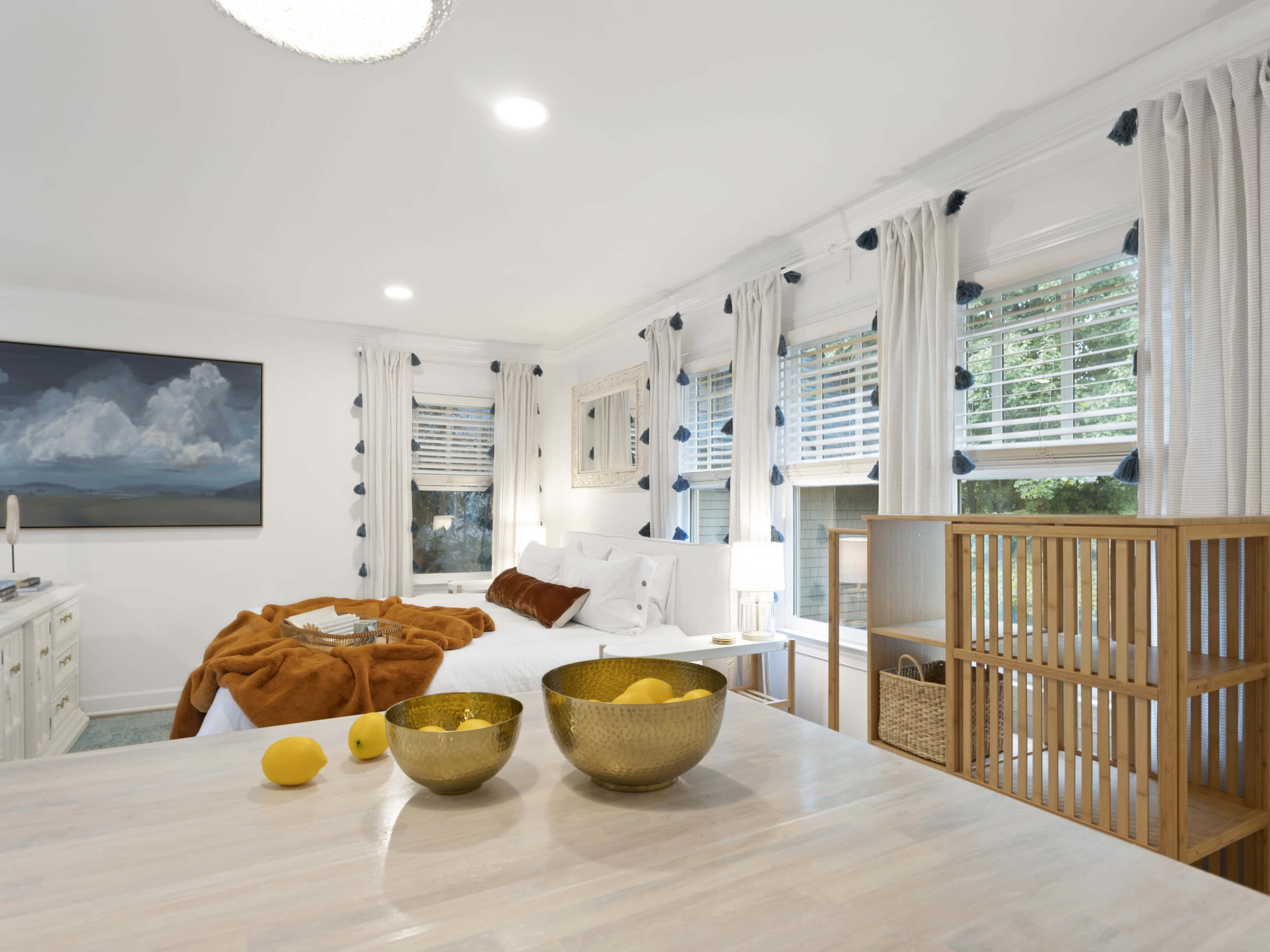 A bright bedroom featuring a large bed, a wooden table with a bowl of lemons, and large windows with white curtains.
