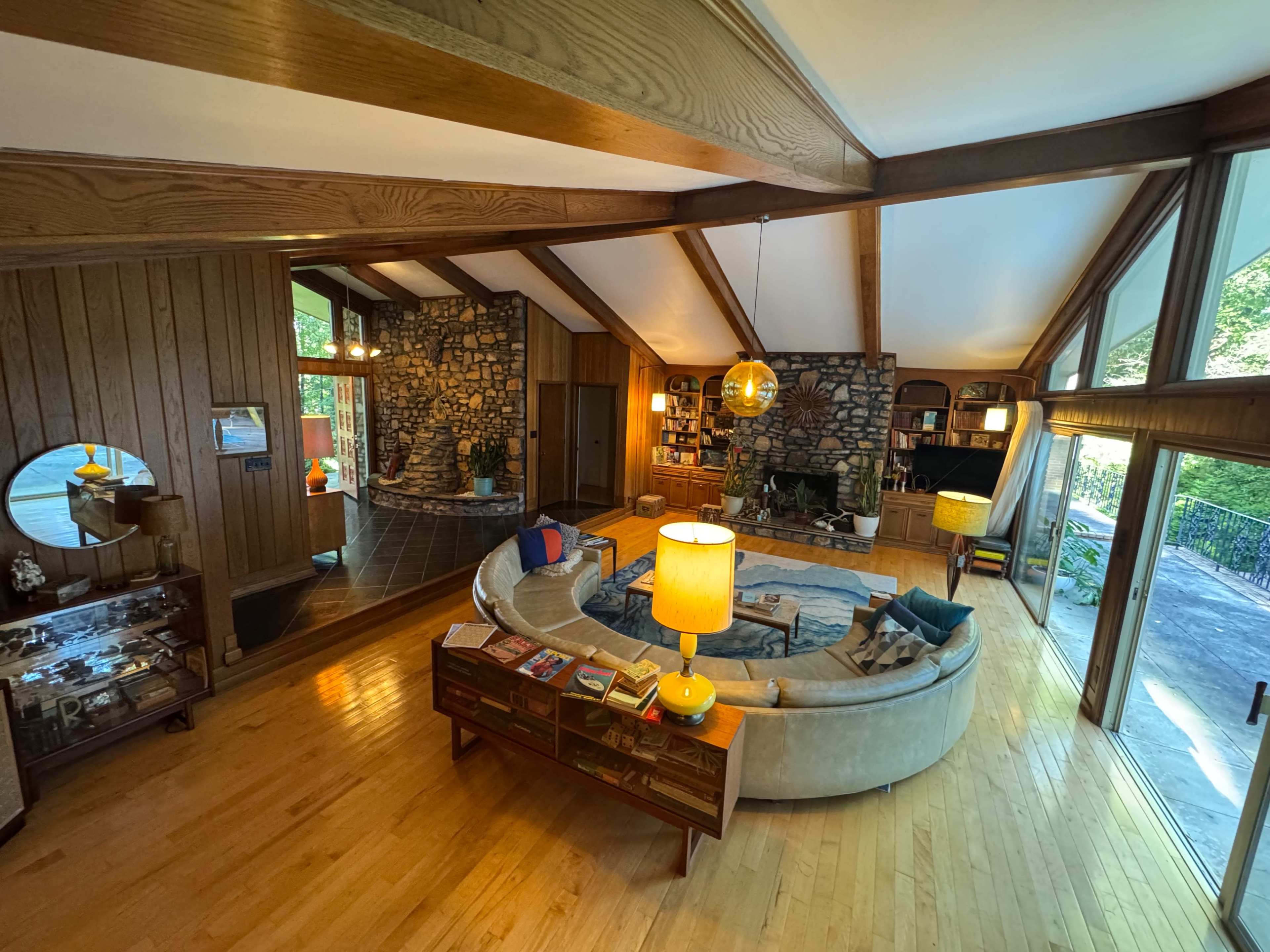 The image shows a spacious living room featuring a circular couch, wooden beams on the ceiling, and a stone wall, with large windows that let in natural light.