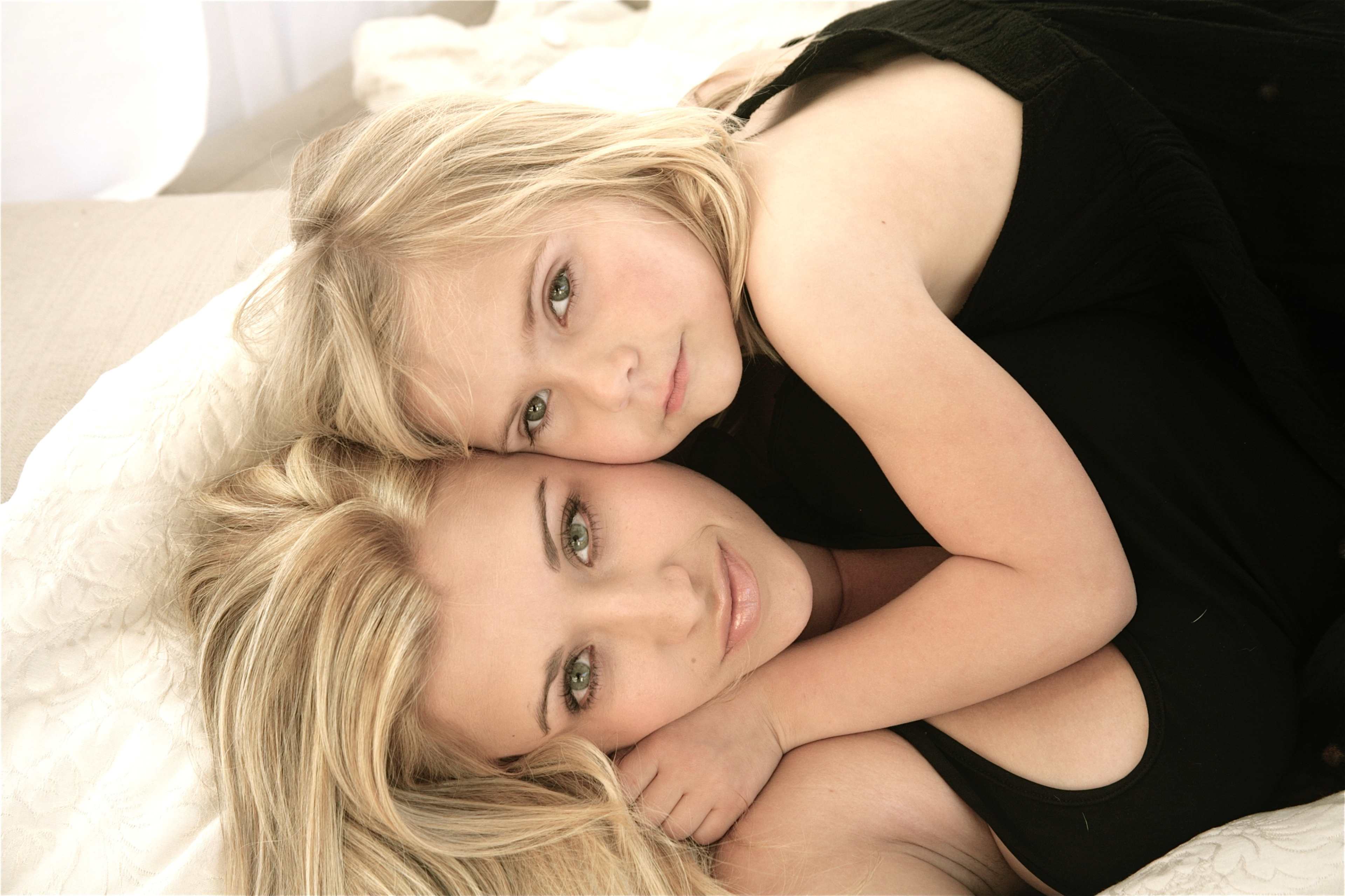A woman and a young girl lie closely together on a light-colored bed, with the girl resting her head on the woman's shoulder.