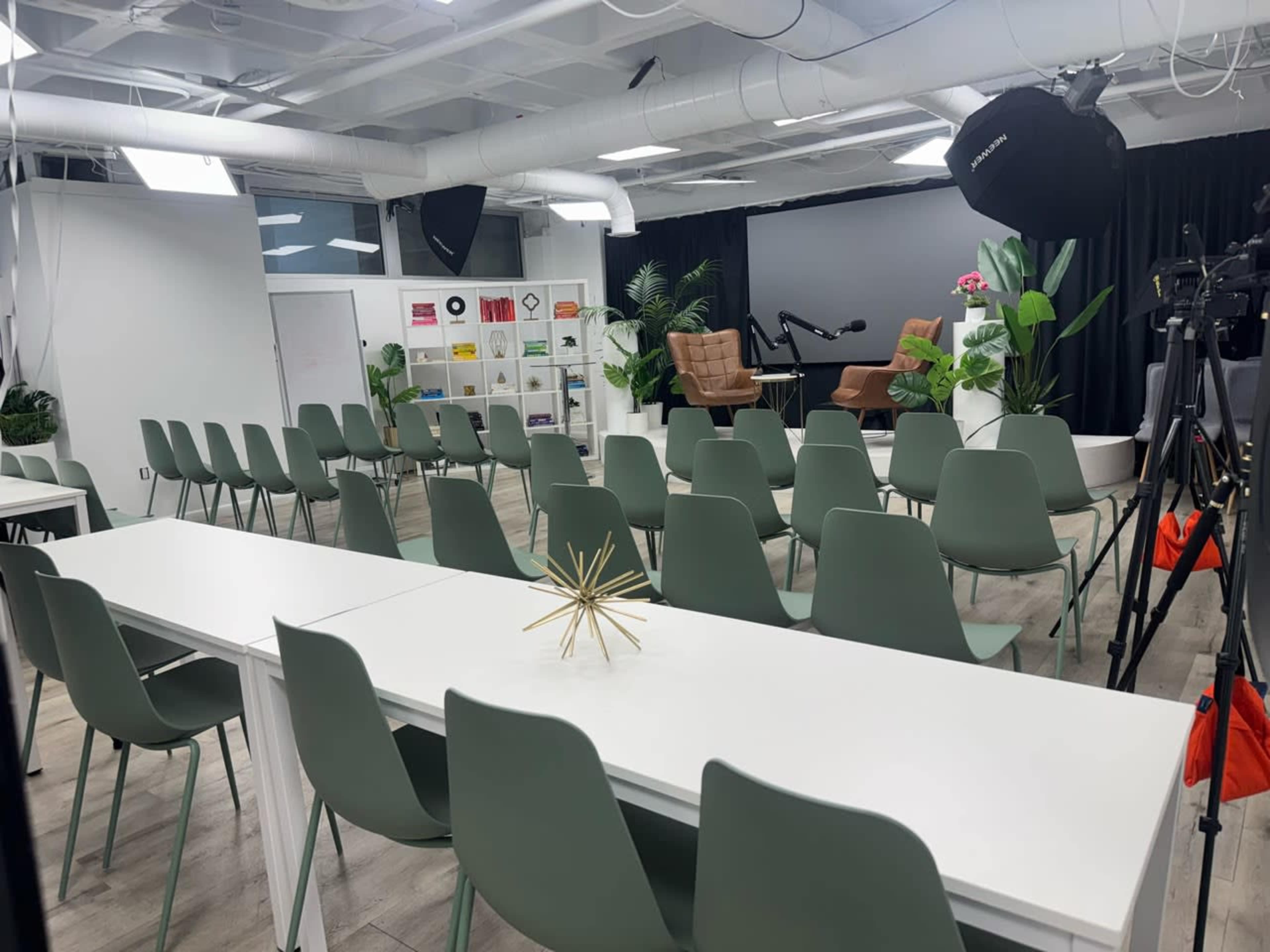 The image shows a modern conference room set up with rows of green chairs facing a stage area that includes two large armchairs and potted plants, with a long white table in the foreground.