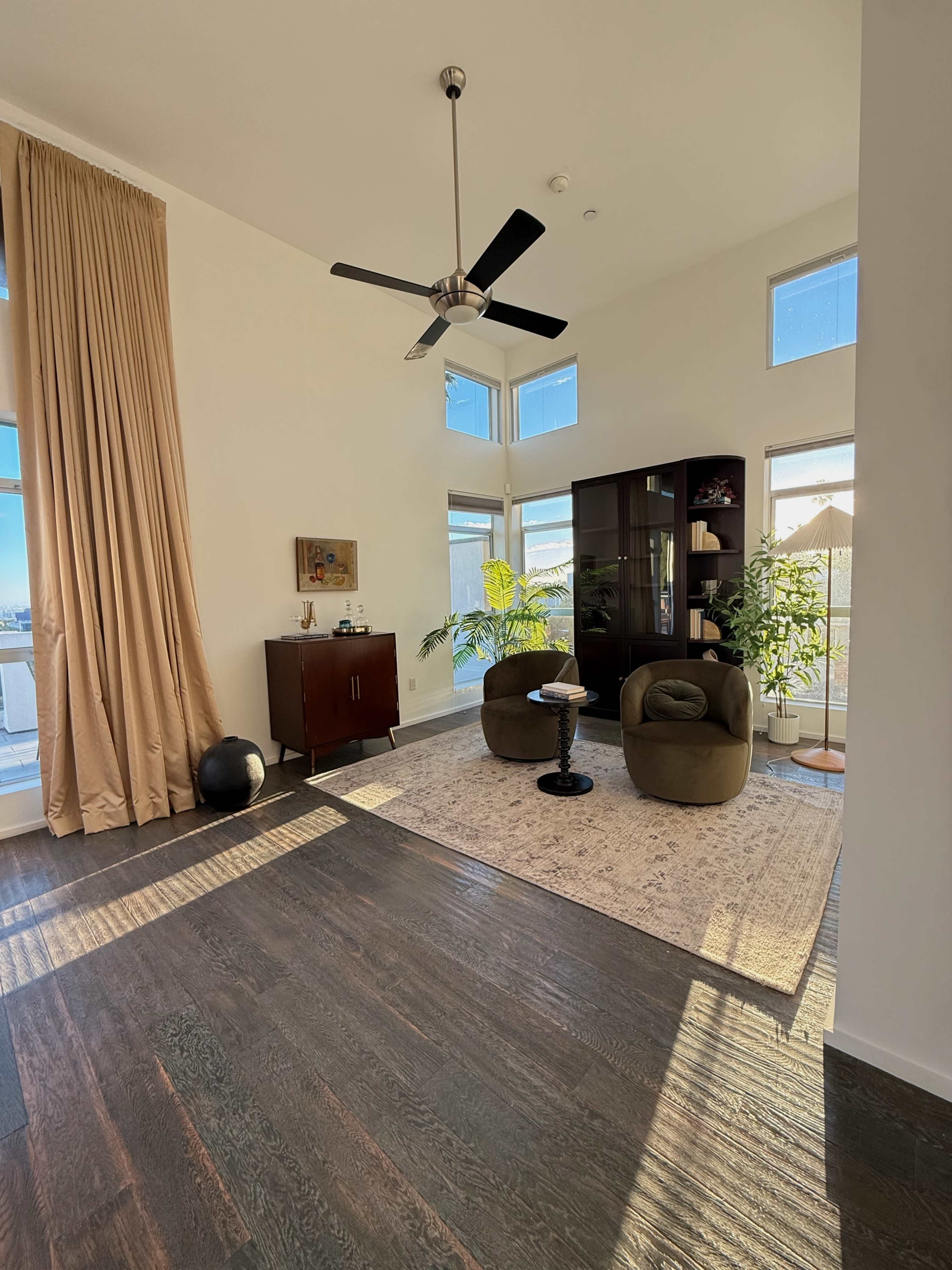 The image shows a bright living space with large windows, a light-colored wall, a dark wooden floor, two green armchairs, and a modern cabinet alongside plants.