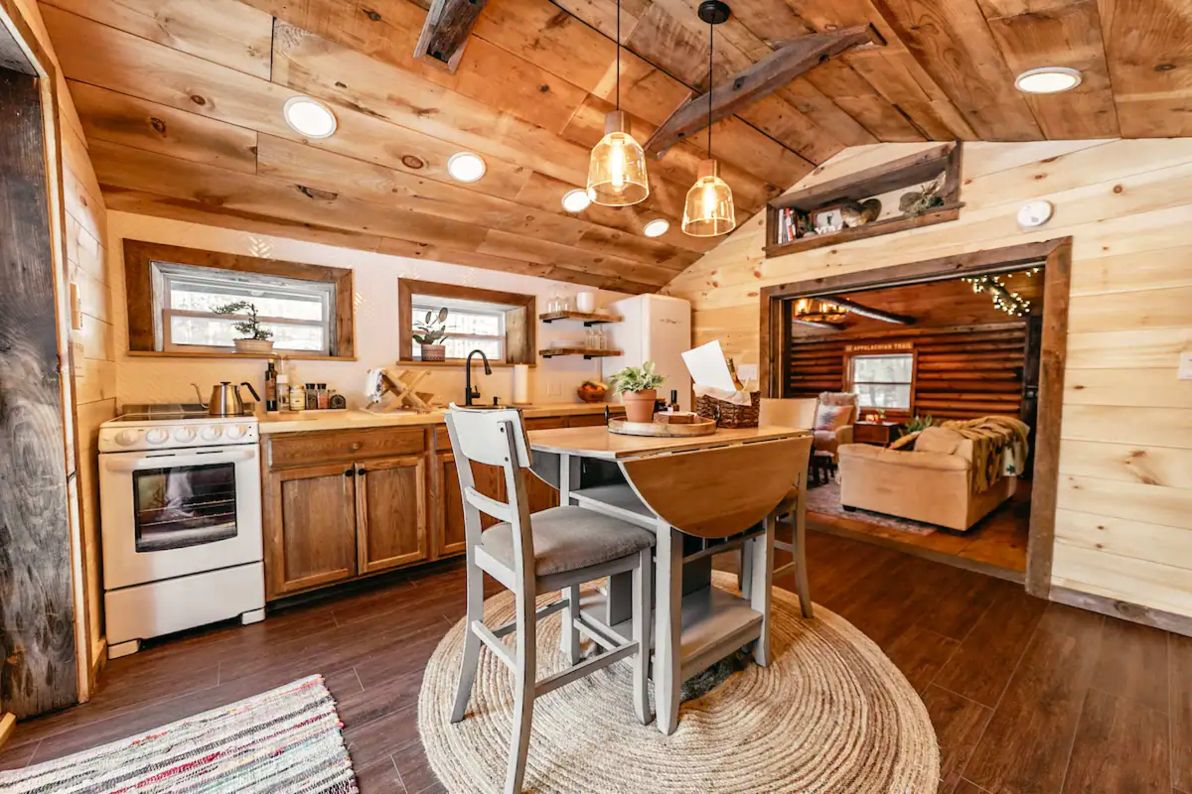 A cozy kitchen with a wooden ceiling, a dining table for two, and a living area visible through an archway.