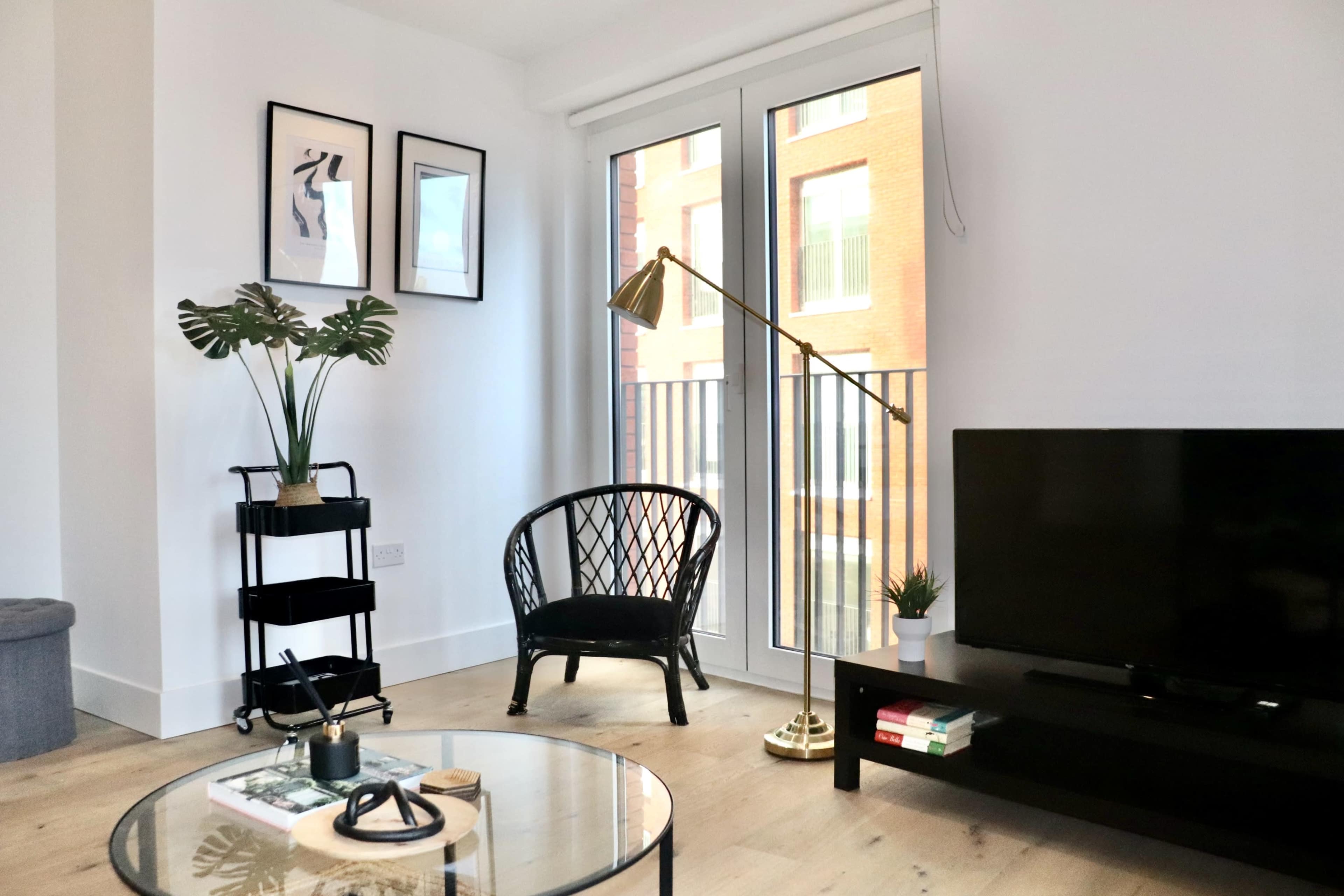 A modern living room with a black chair, a glass coffee table, a TV stand, potted plants, and large windows overlooking a balcony.