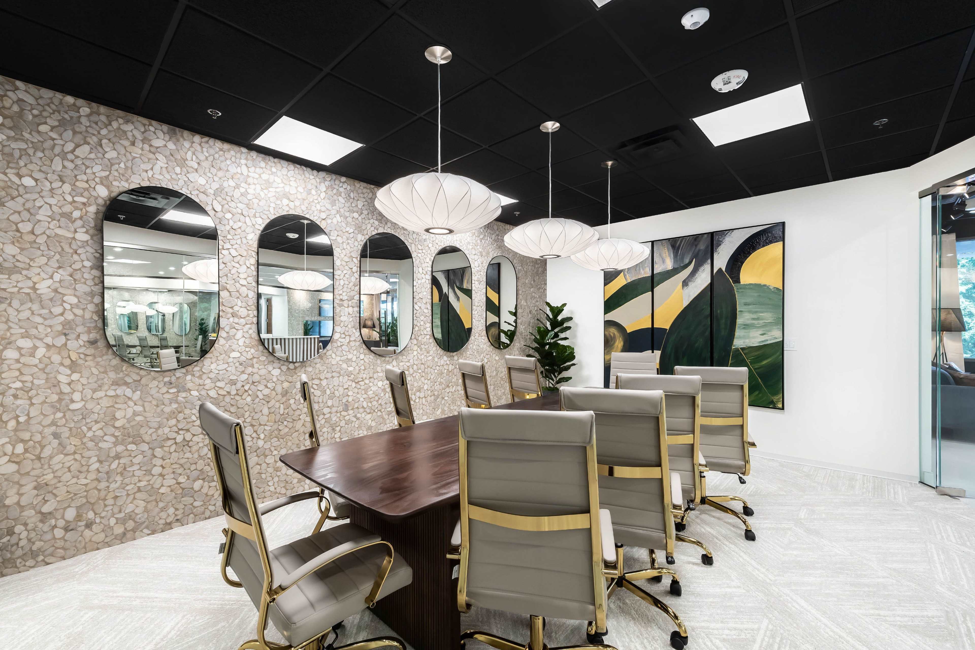 A modern conference room features a long wooden table surrounded by gray leather chairs, with a textured stone wall and several round mirrors, illuminated by pendant lights.
