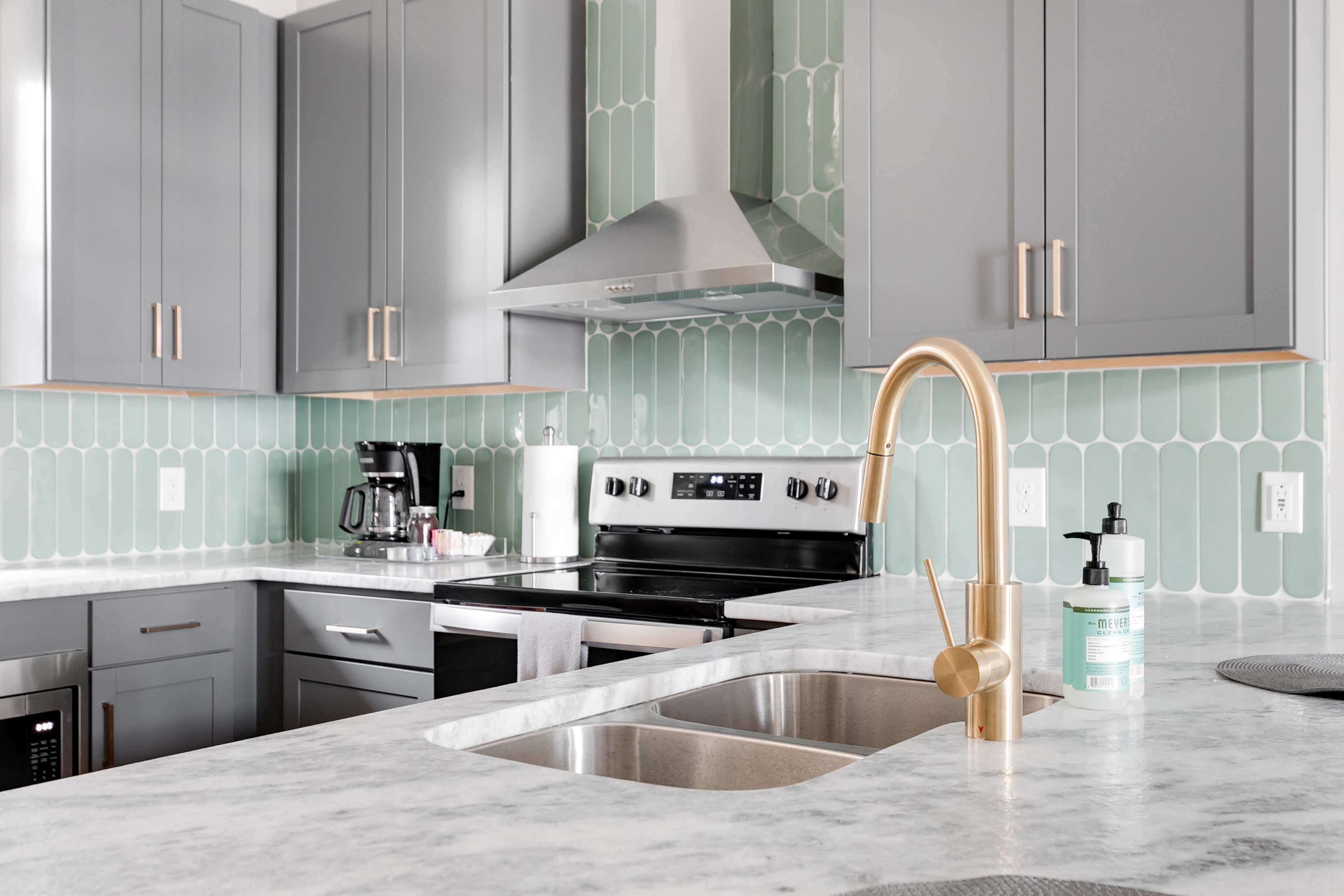 A modern kitchen features gray cabinets, a marble countertop with a double sink, and green tile backsplash.